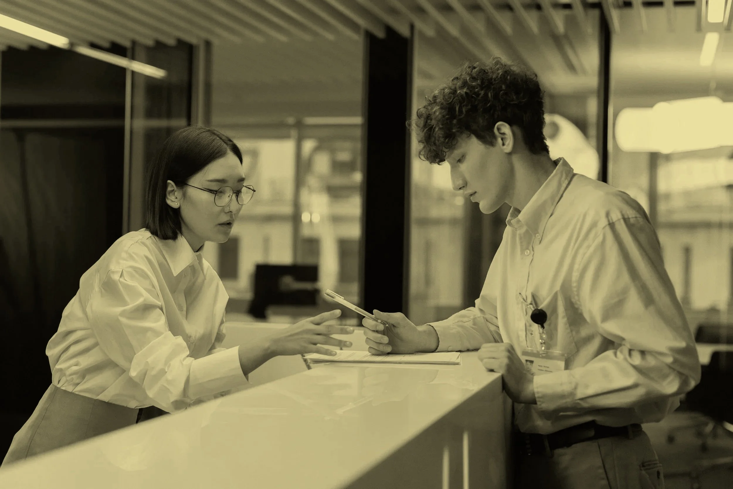 Two young adults, a woman with glasses and a man with curly hair, are working together at a counter, looking at a smartphone, in what appears to be a modern office or laboratory setting.