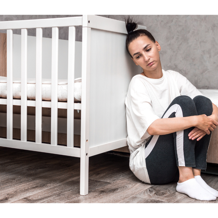 New mother sitting on floor hugging her knees with back against crib. She looks tired and overwhelmed.