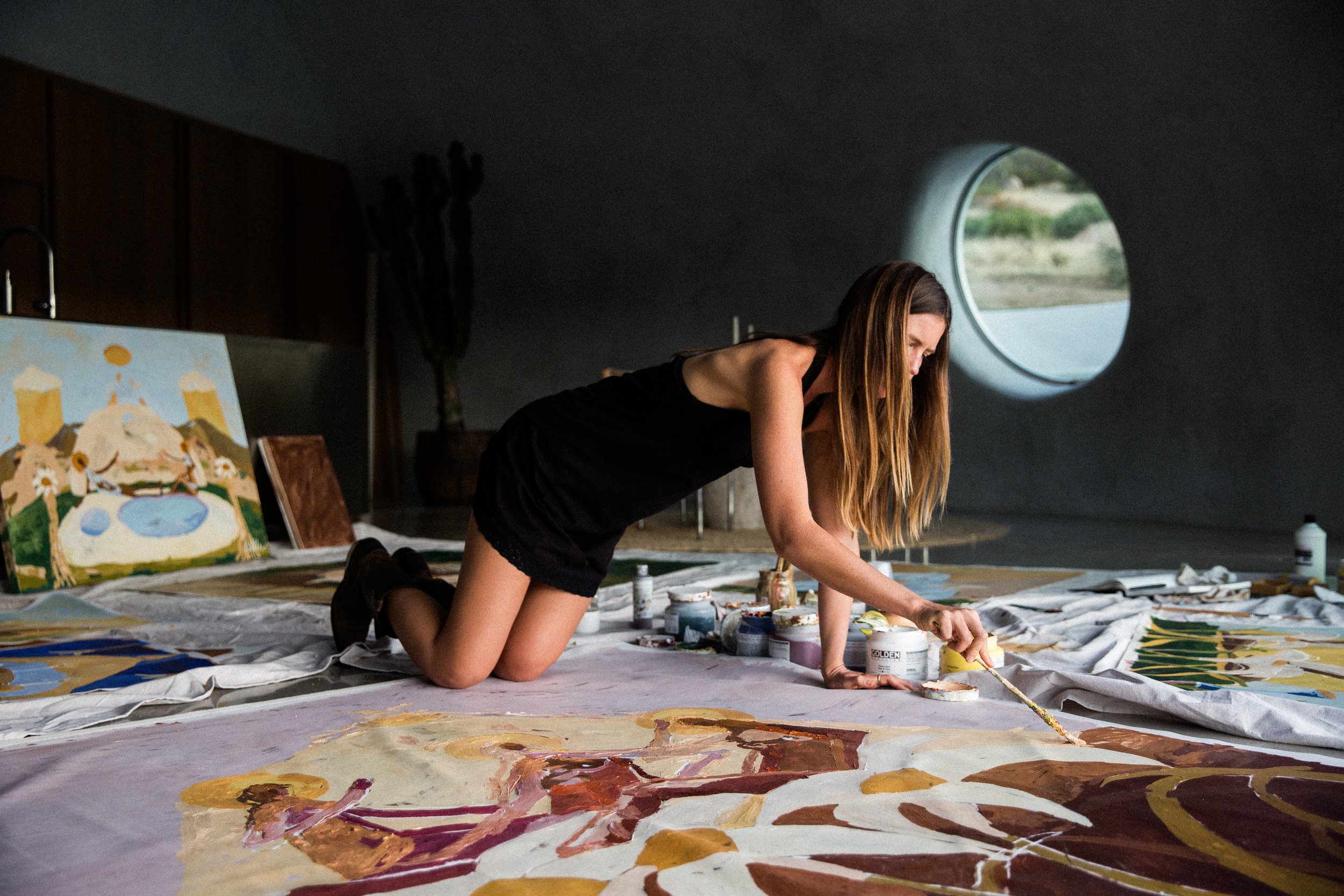 A woman kneeling on the floor in an art studio, painting a large canvas with acrylic paints, surrounded by other paintings and art supplies in a dimly lit room with a round window.