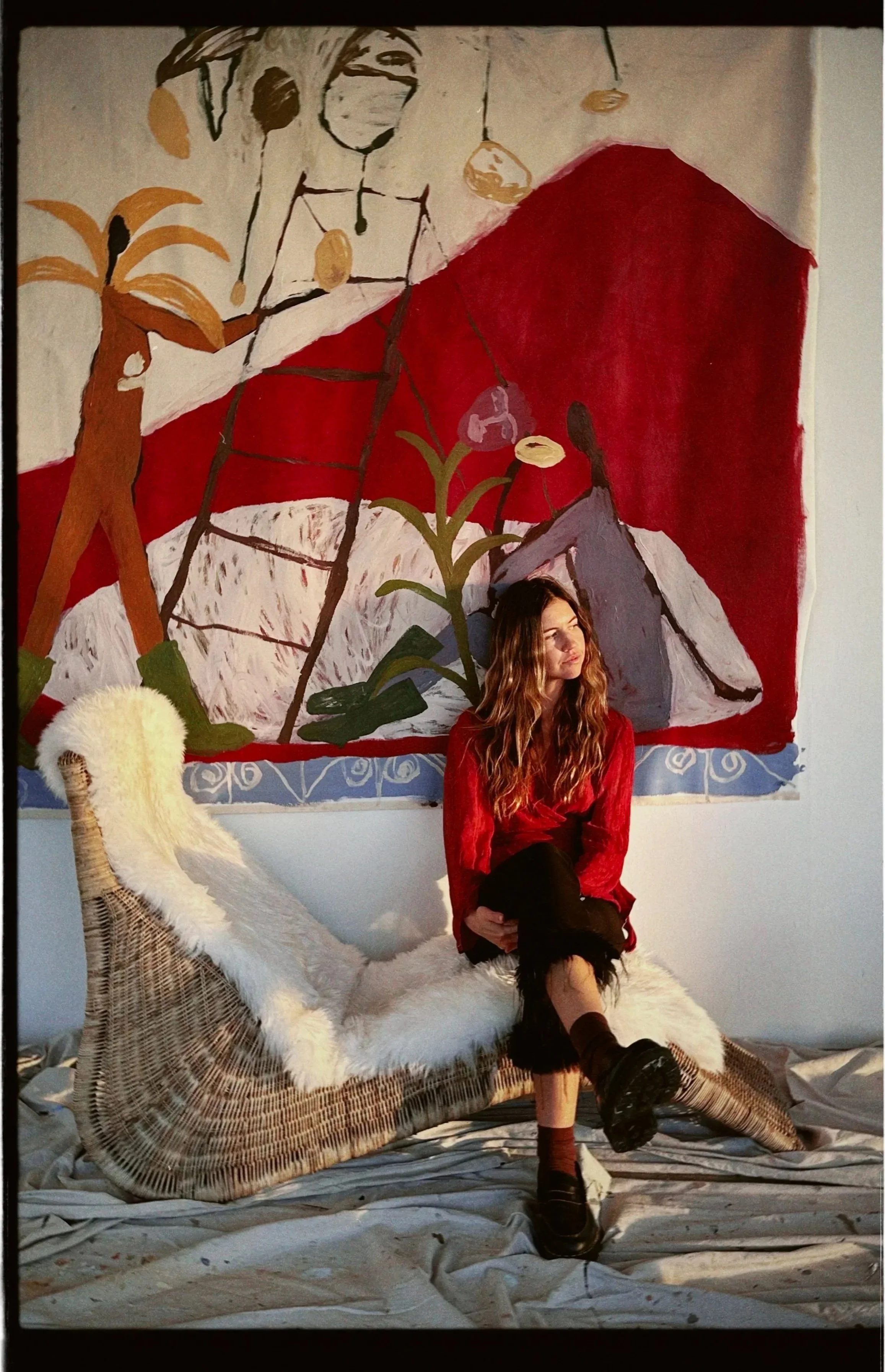 A woman with long wavy hair, wearing a red shirt and black pants, sitting on a wicker chaise lounge with a white furry cover, in front of an abstract mural with red, white, and brown colors and diverse shapes, including plants and geometric forms.