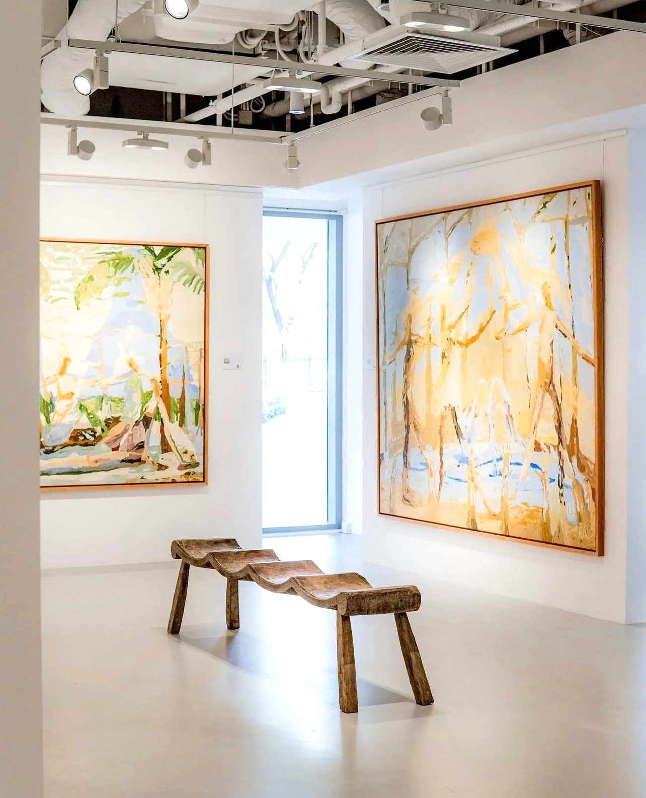 Art gallery with two large colorful abstract paintings on white walls, a wooden bench in the center, and a glass door letting in natural light.