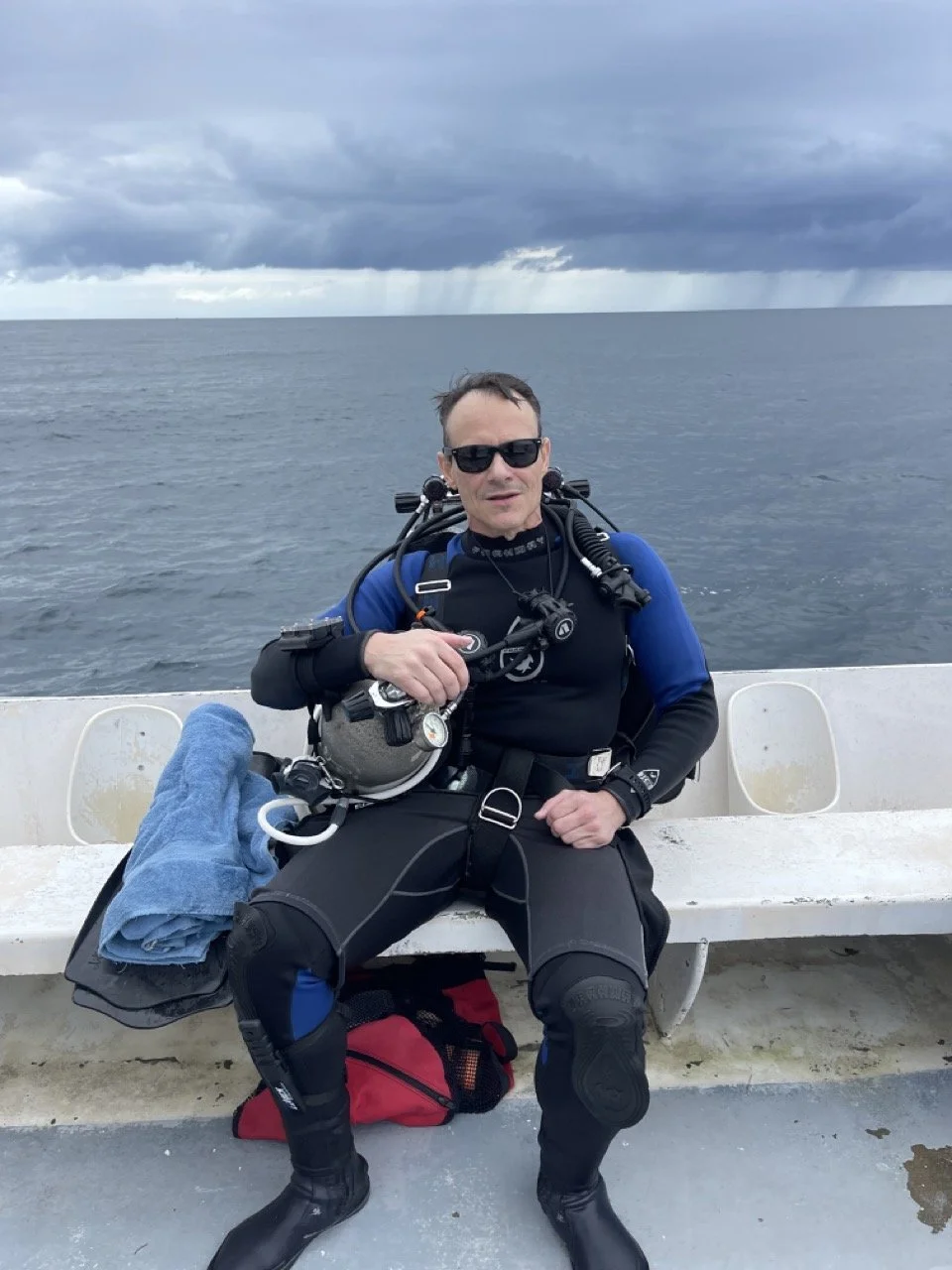 Scuba diver in a wetsuit sitting on a boat with diving gear under cloudy skies.