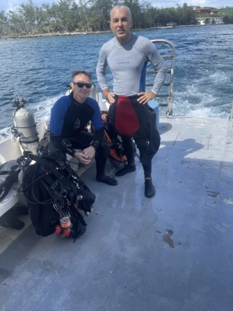 Two people in wetsuits on a boat with scuba diving gear, ocean and trees in background.