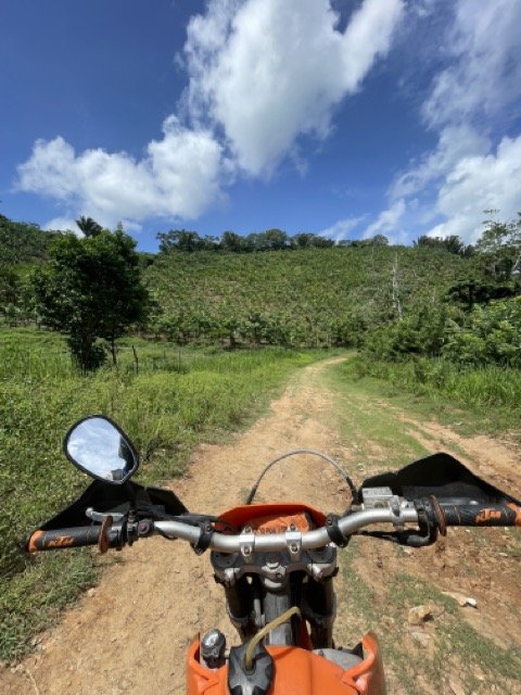 View from a motorcycle handlebar on a dirt road through a green landscape with hills under a blue sky with clouds.