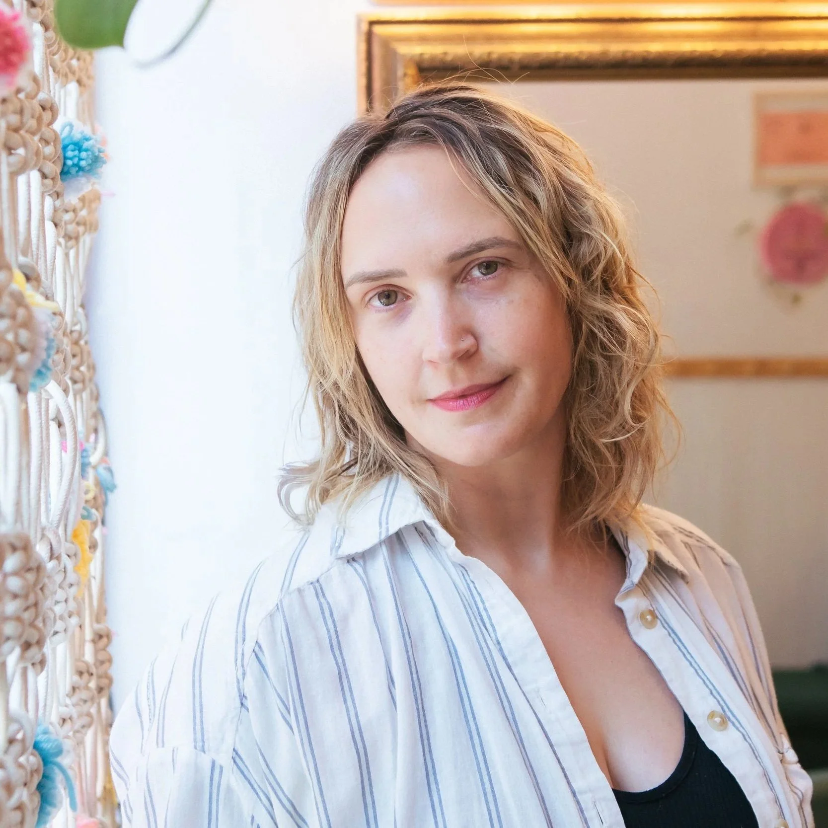 A woman with short blonde hair and fair skin, wearing a white top and brown cardigan, standing in a room with floral wallpaper, a wooden shelf with plants and bottles, and a mirror.