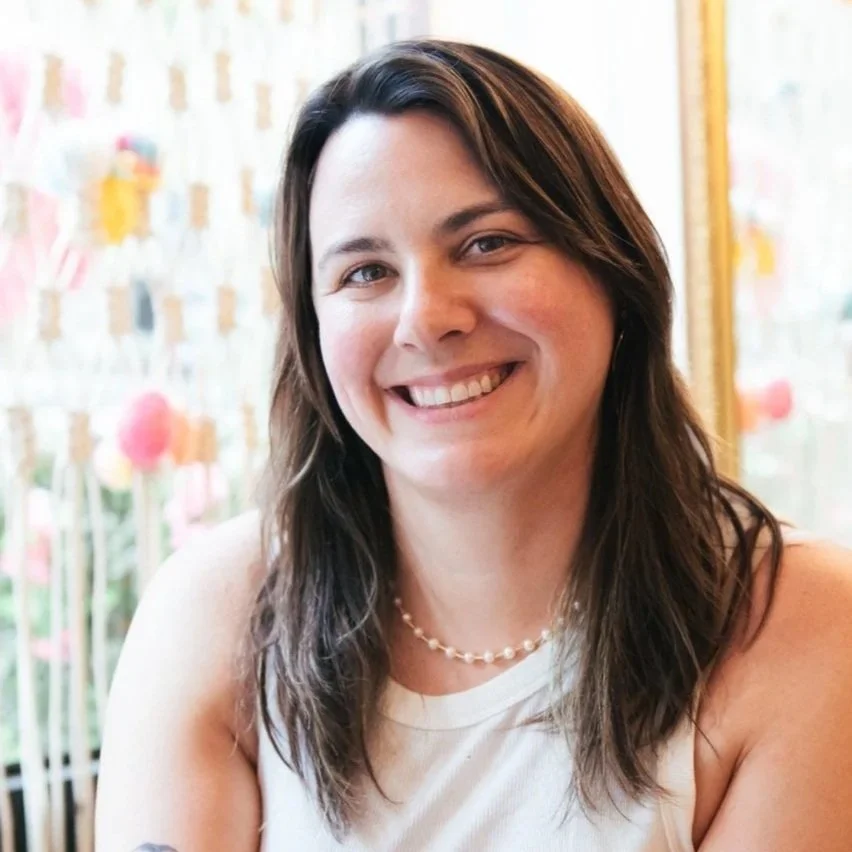 A woman with brown hair wearing a white t-shirt and gold jewelry, standing in front of a decorative backdrop made of woven rope and colorful yarn and embellishments, with a pink fringed decoration behind her.