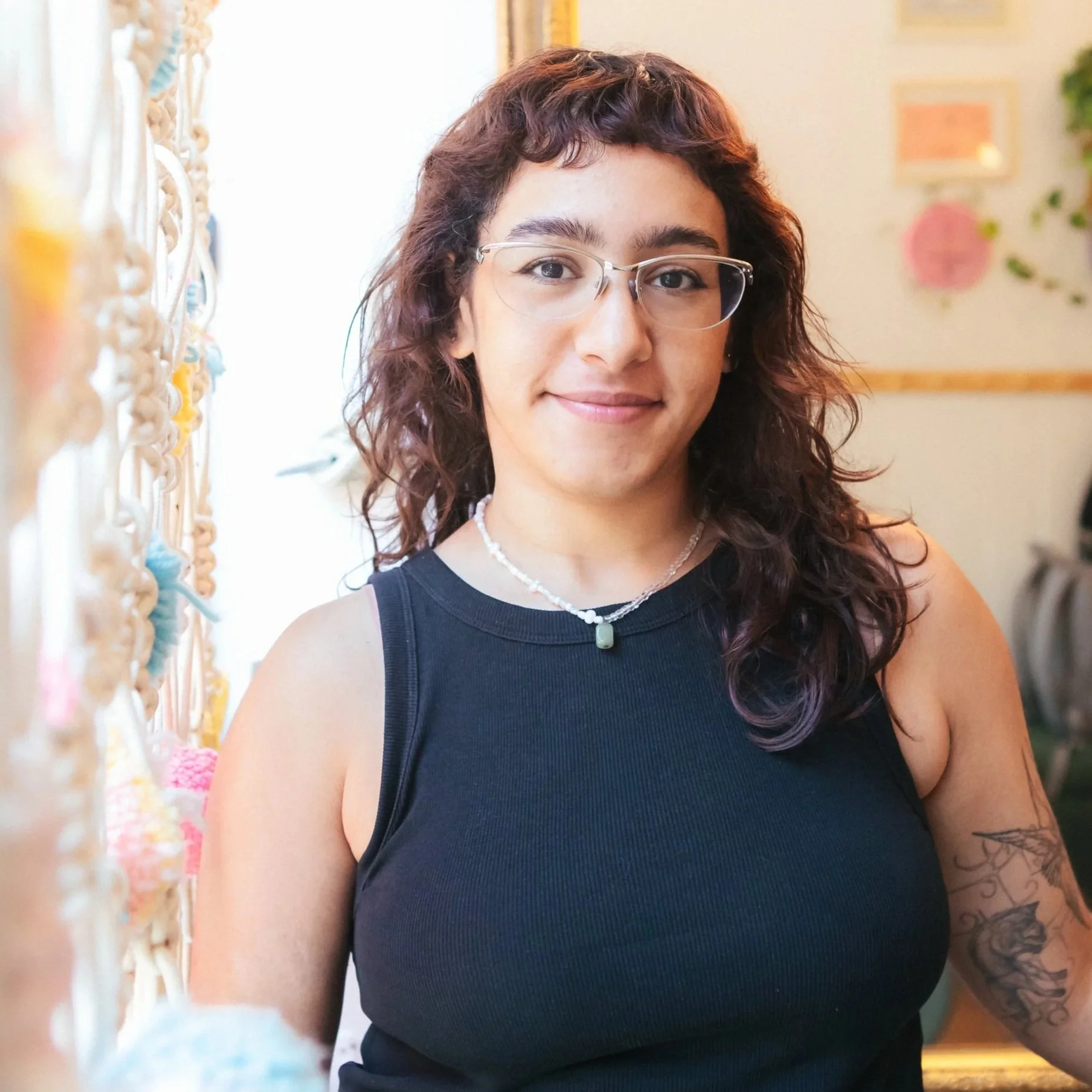 A woman with wavy red hair and glasses, smiling, wearing a white t-shirt and gold jewelry, in a cozy indoor setting.