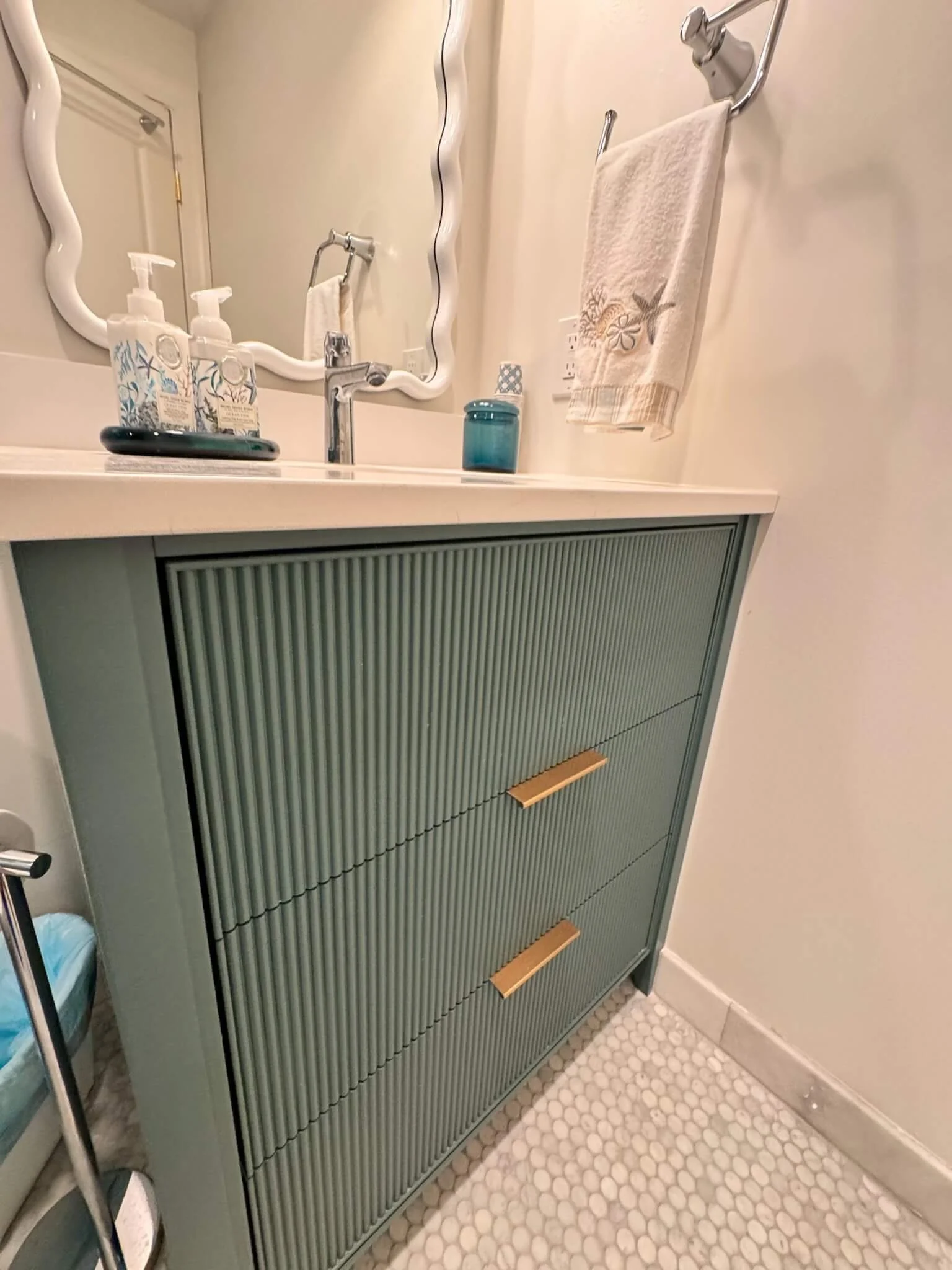 Built-in vanity with LED lighted round mirror, marble tile walls, and gold hardware in an Ocean City spa-inspired bathroom by 2B Contracting.
