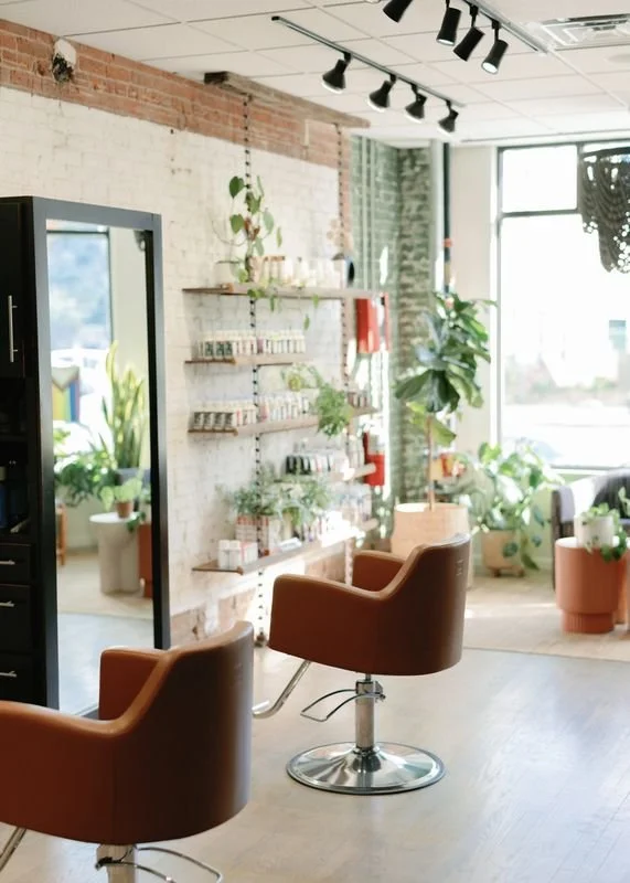Interior view of a hair salon or spa with barber chairs, shelves with hair products, and large windows with plants.