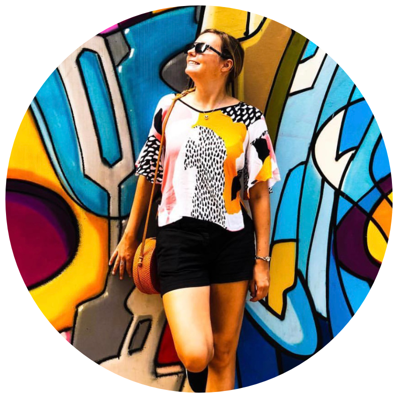 Polly Taylor smiling and wearing sunglasses, dressed in a colourful, abstract-patterned top and black shorts, standing in front of a vibrant graffiti wall.