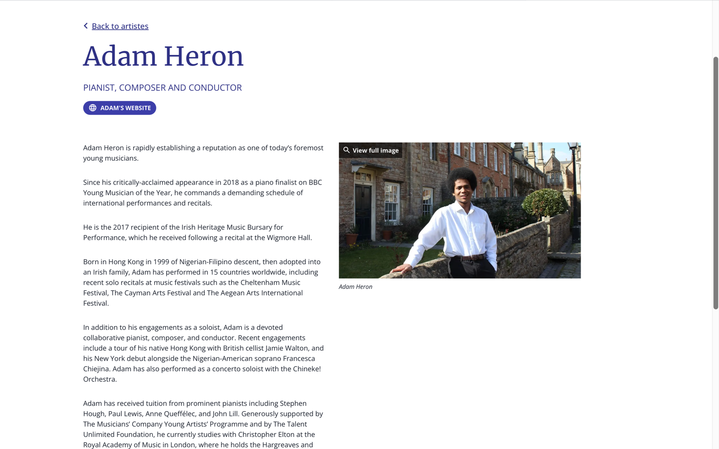 Portrait of Adam Heron, a young man with curly black hair, wearing a white shirt, standing outdoors near a stone wall with buildings in the background.