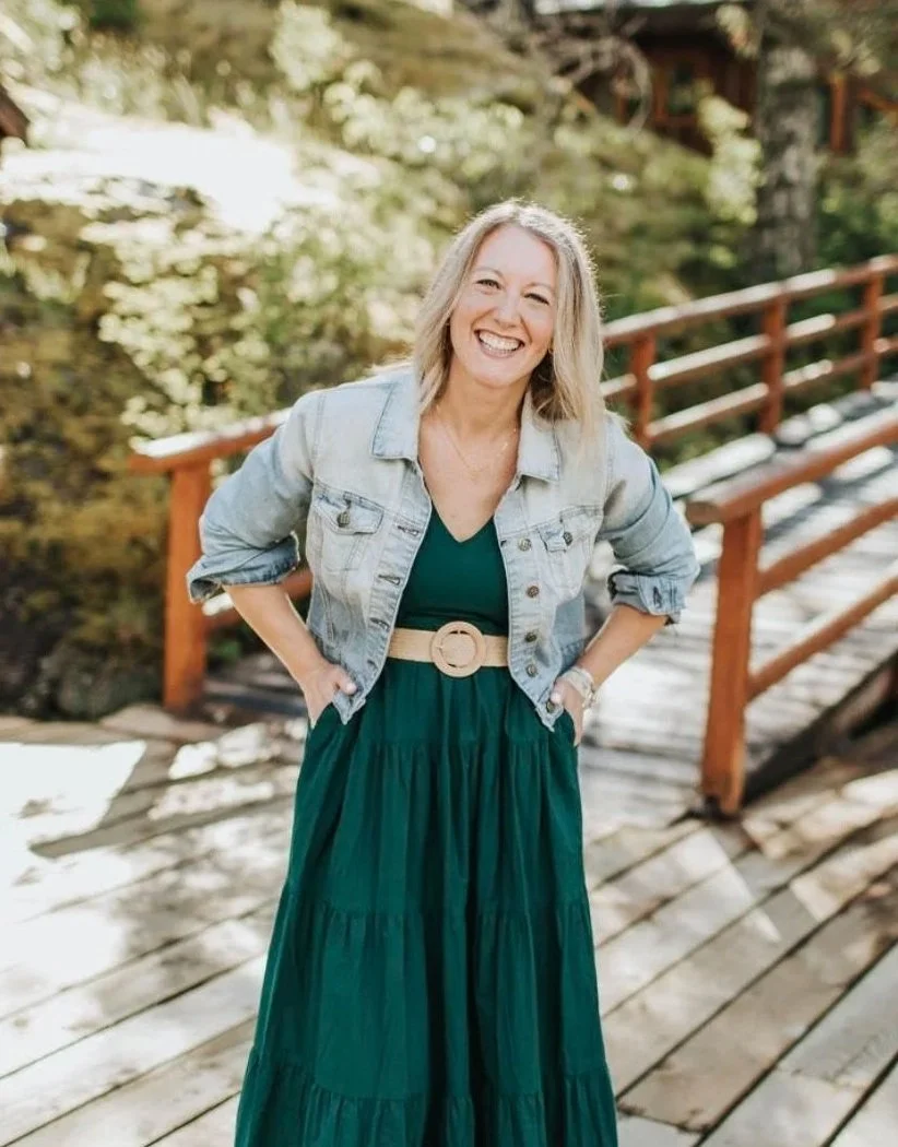 A woman with blonde hair wearing a denim jacket and a long green dress with a beige belt, smiling and standing outdoors on a wooden bridge surrounded by greenery.