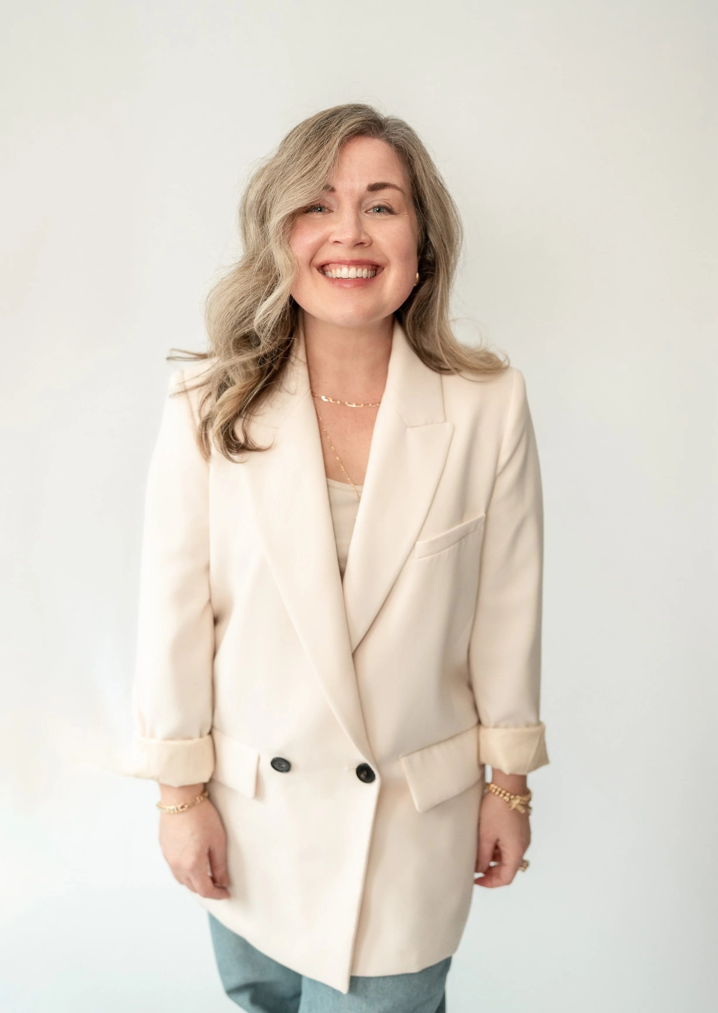 A smiling woman with wavy blonde hair wearing a cream-colored blazer, gold jewelry, and light blue jeans standing against a plain white background.