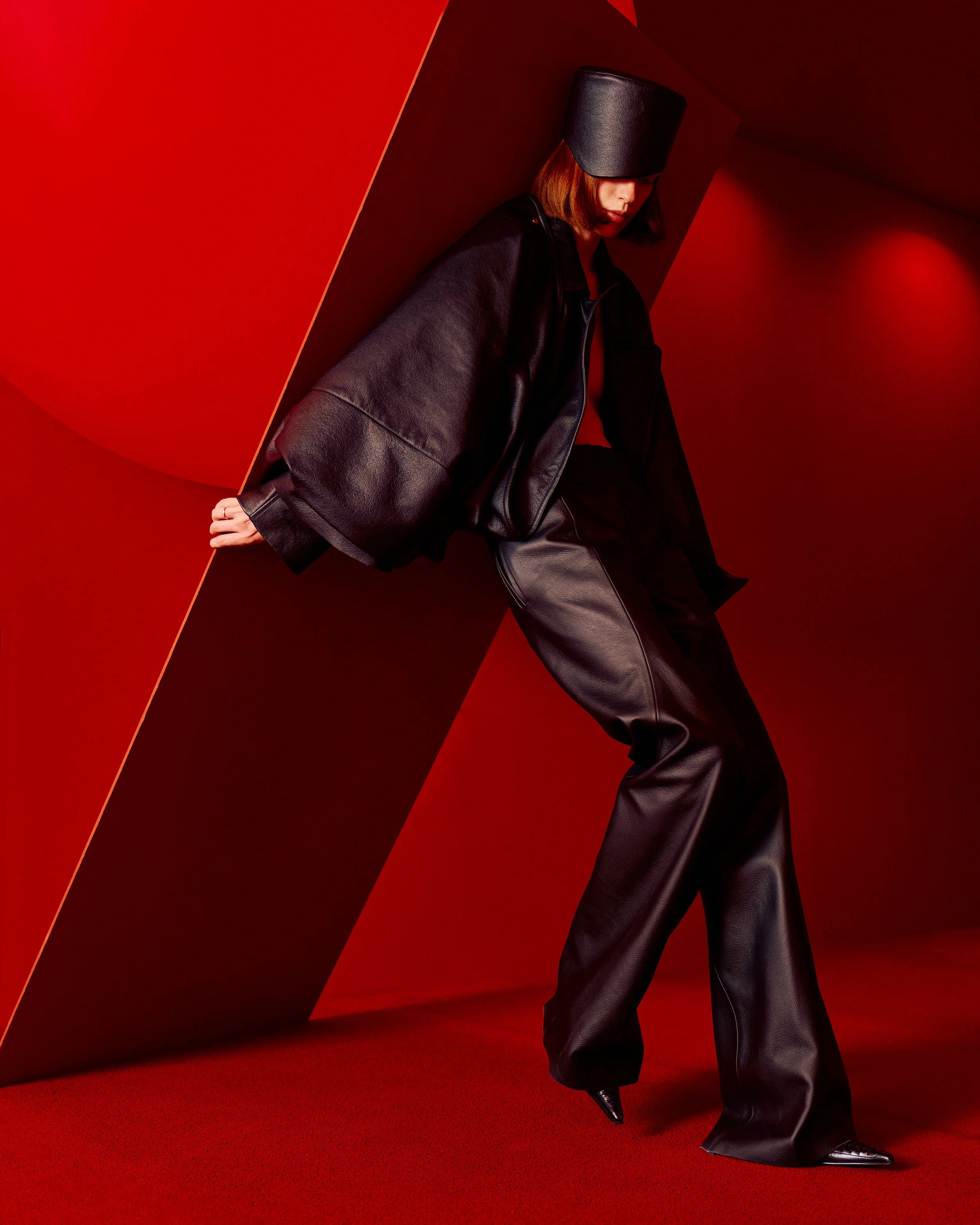 Woman dressed in black leather jacket and bag, wearing black leather pants and shiny black shoes, posed against red background with angular shapes, with her head covered by a large black hat.