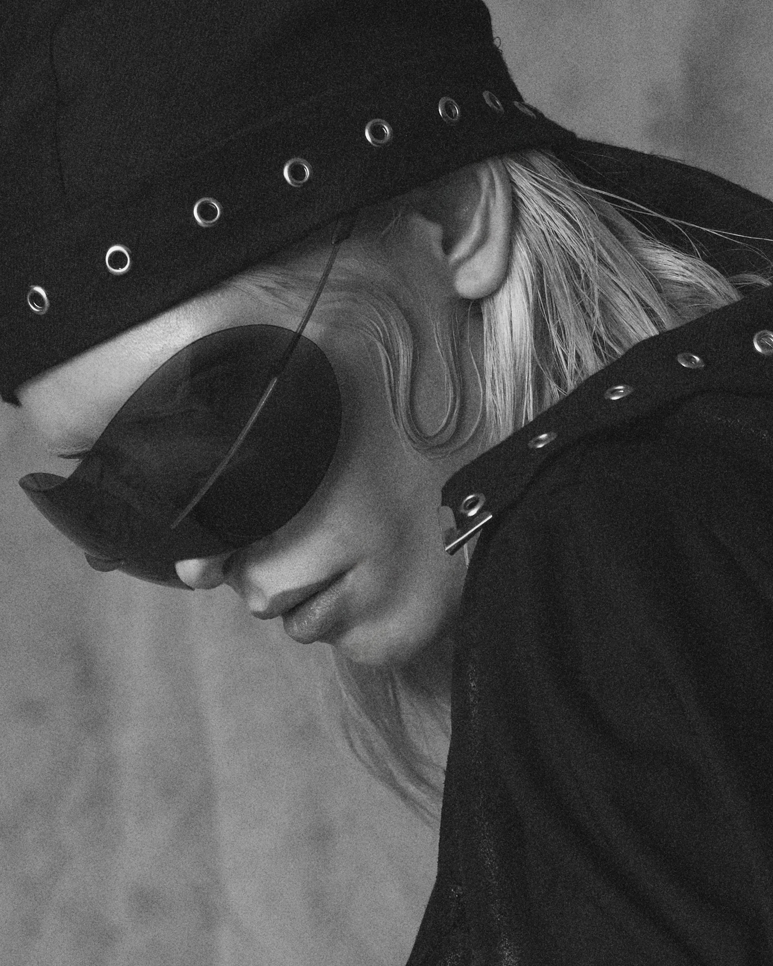 Black and white close-up of a person's face with their eyes closed, wearing a black hat with eyelets, and holding a long, thin object near their eye.
