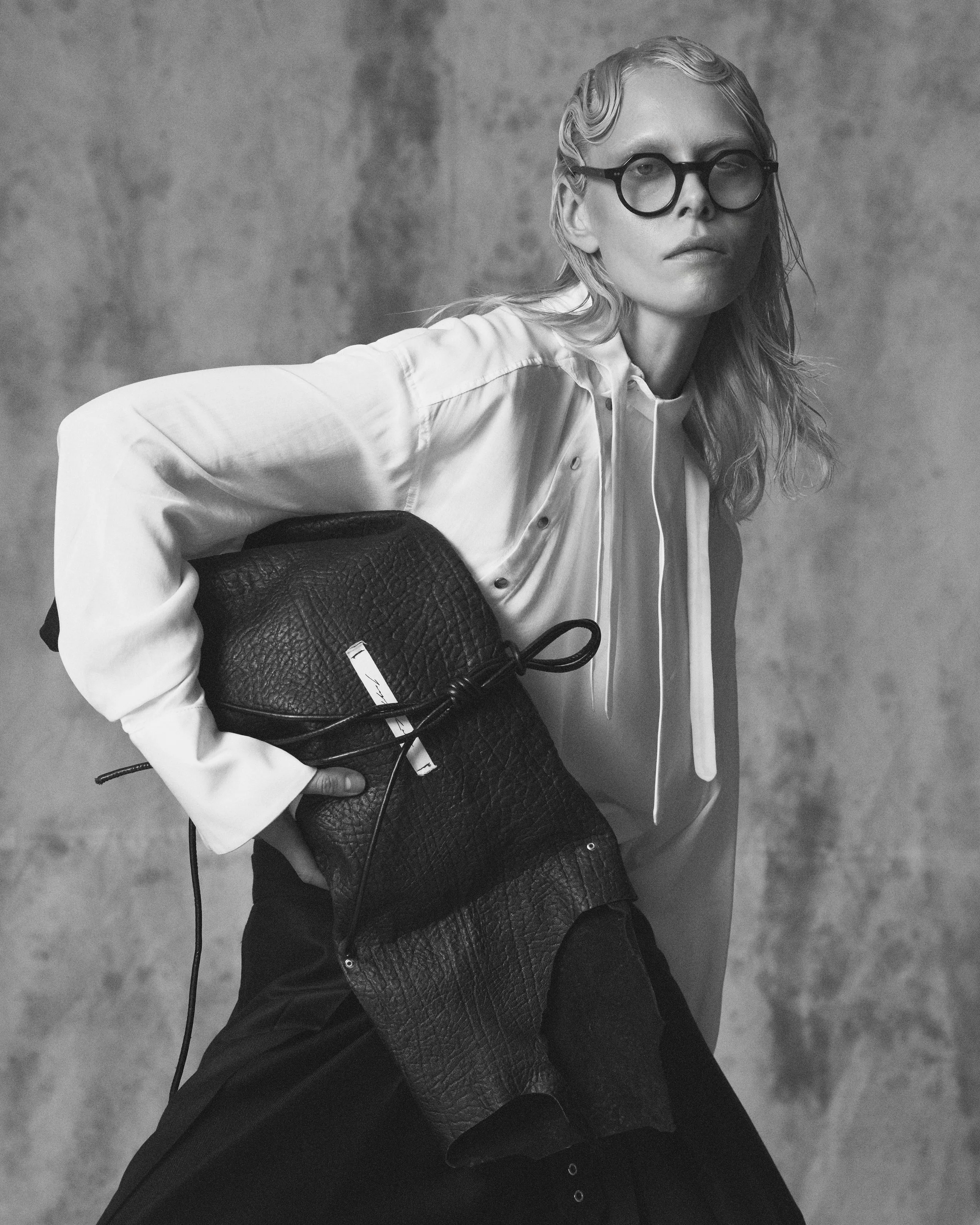 Black and white photo of a woman with wavy hair wearing large round glasses, a light-colored button-up shirt, and black pants, holding a textured black bag, against a plain background.