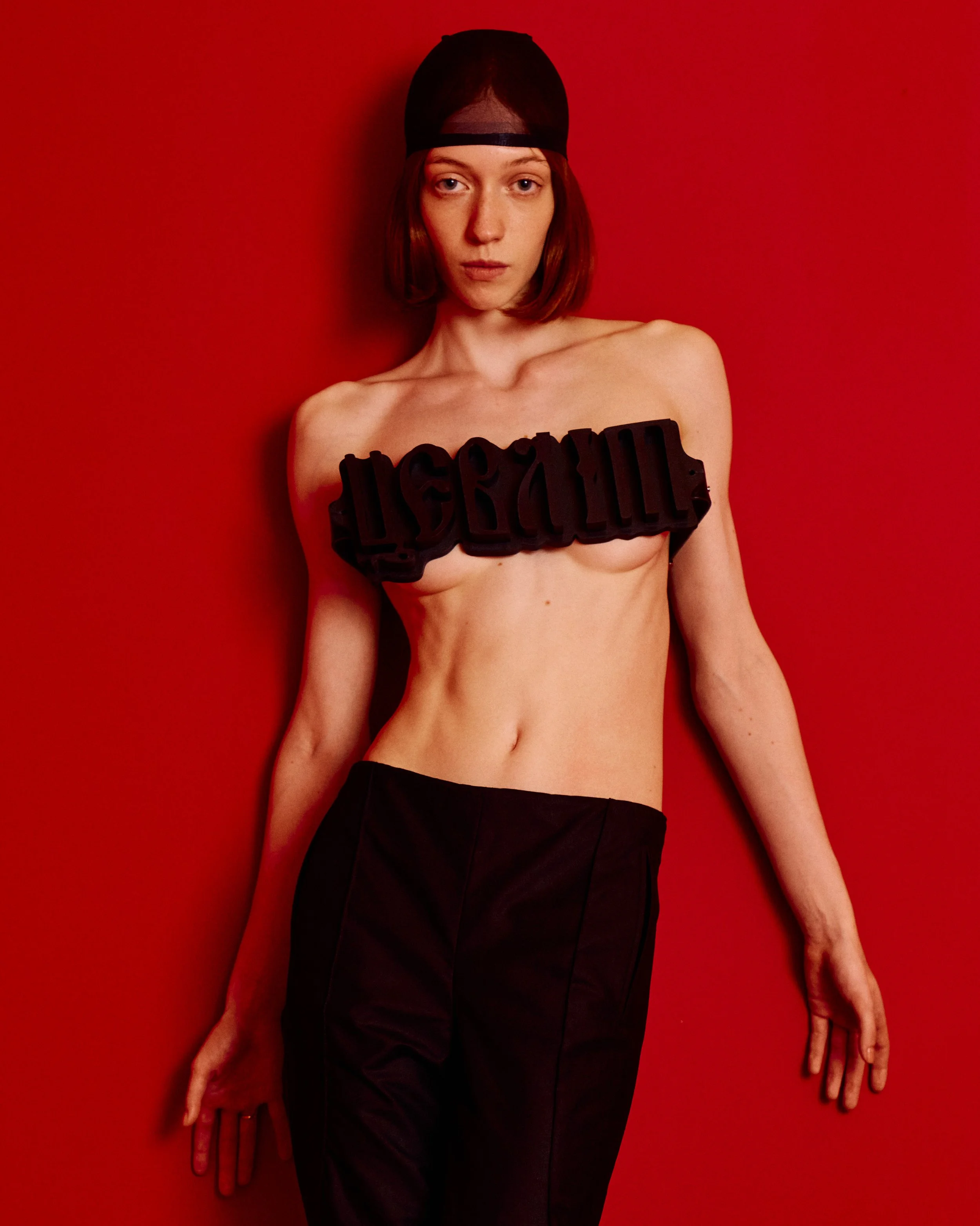 A woman with short brown hair stands against a red wall, wearing a black mesh head covering, black pants, and a black foam sign with unreadable text across her upper chest, with her arms extended downward and hands relaxed.