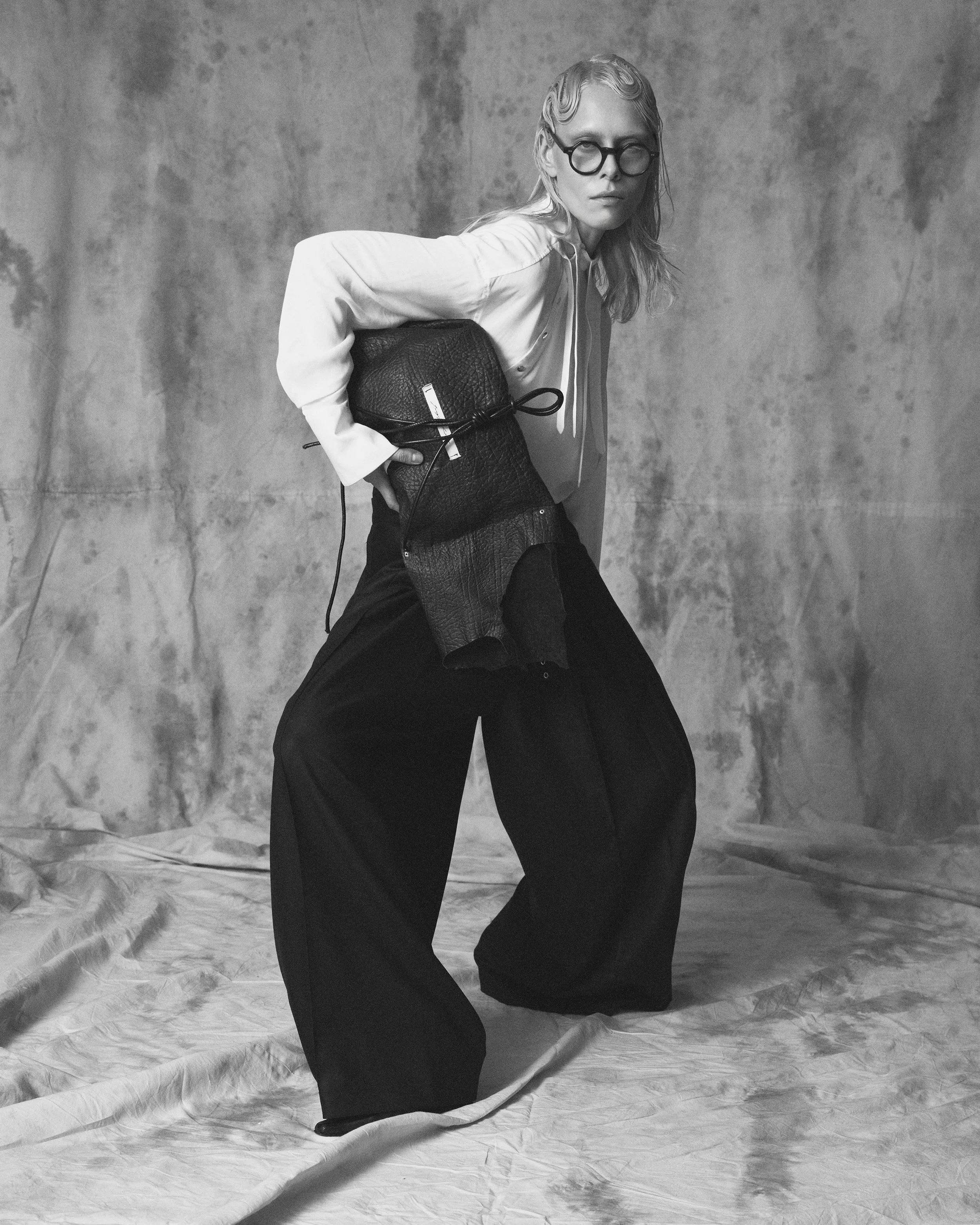 Black and white photo of a woman with wavy hair and glasses, wearing a satin blouse and black wide-leg pants, holding a textured handbag, standing against a plain textured wall, with her head slightly tilted and looking directly at the camera.