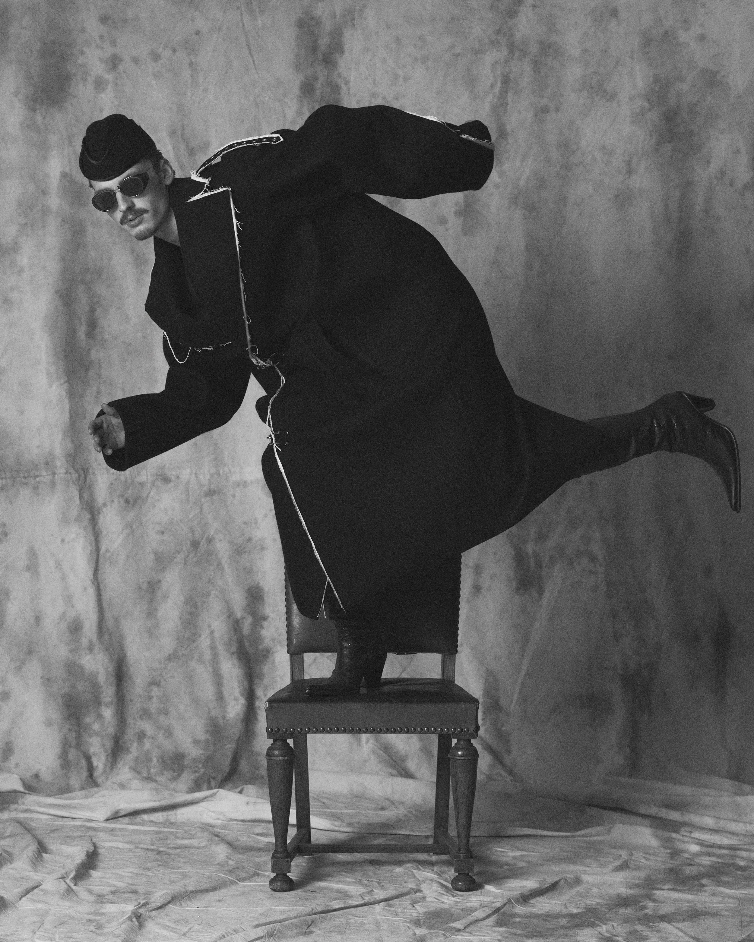 Black and white photograph of a man dressed in dark clothing, wearing sunglasses and a cap, sitting on a wooden chair. The man is leaning forward with his elbow resting on his knee, and his hand touching the back of his head. The background appears t