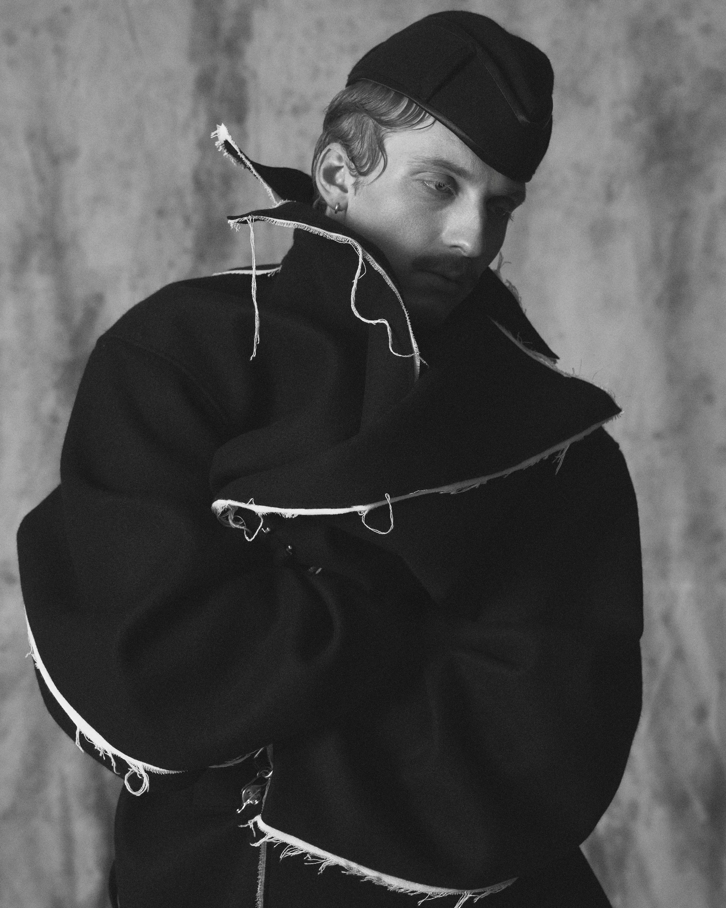 Black and white photo of a young person with short hair, wearing a black beanie and a distressed black jacket with frayed edges, looking down with a somber expression.