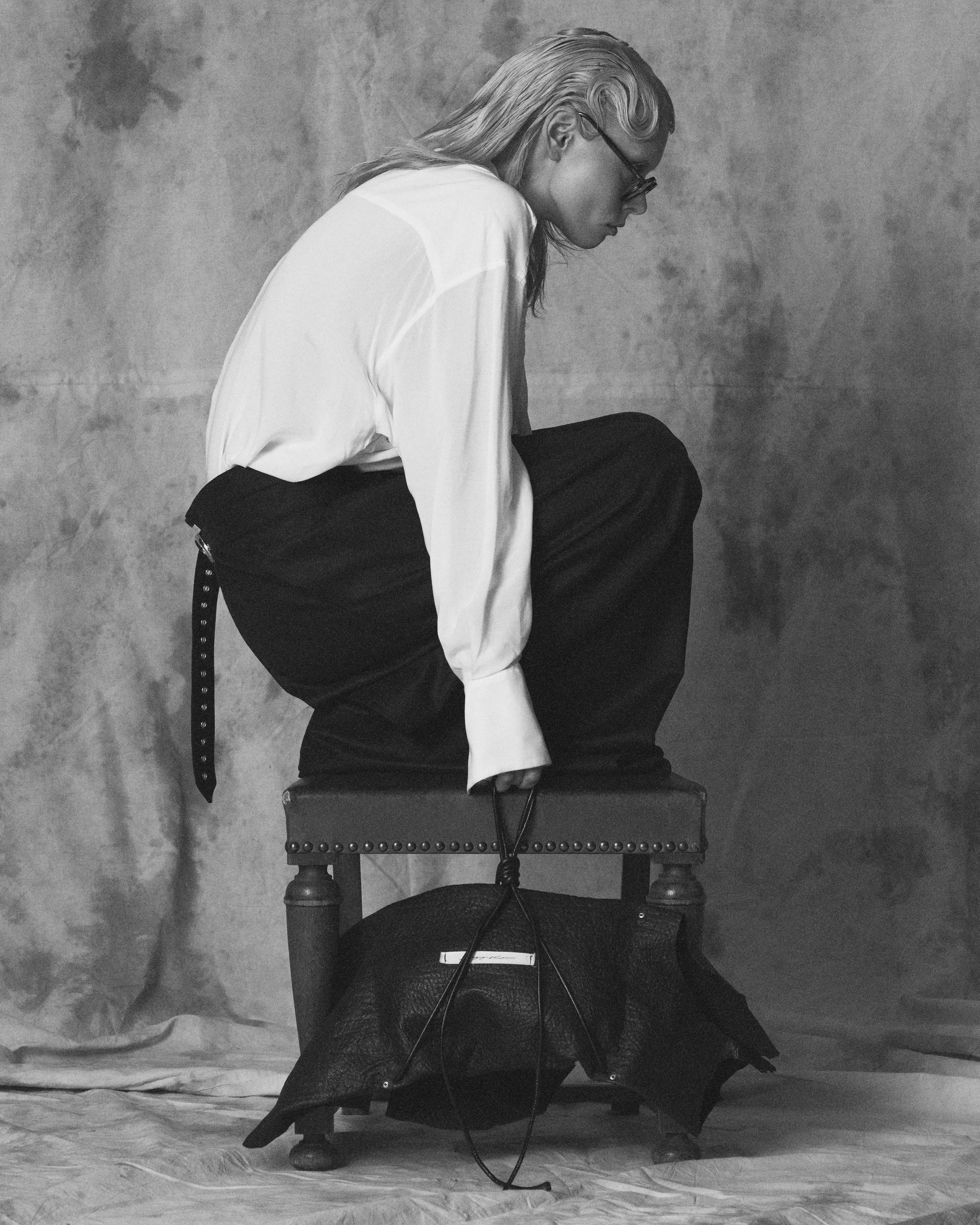A woman with long hair wearing glasses, dressed in a white shirt and dark pants, weeping and sitting on an old wooden chair in front of a textured wall, with her head bent down.