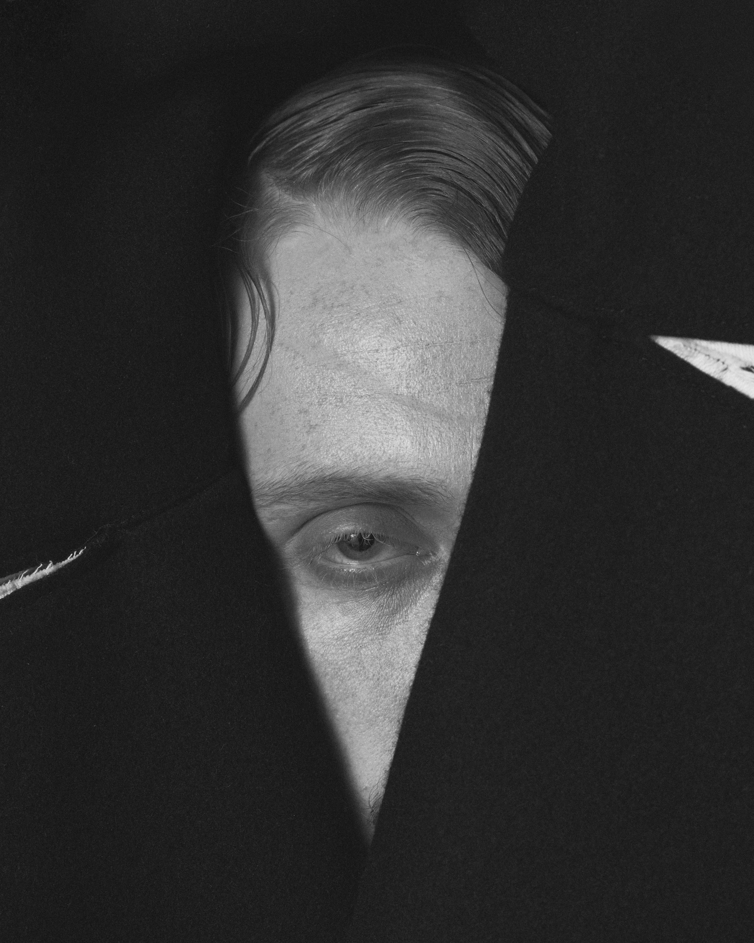 Black and white photo of a person's face, with part of the face obscured by black fabric or shadow, showing one eye and forehead with hair slicked back.