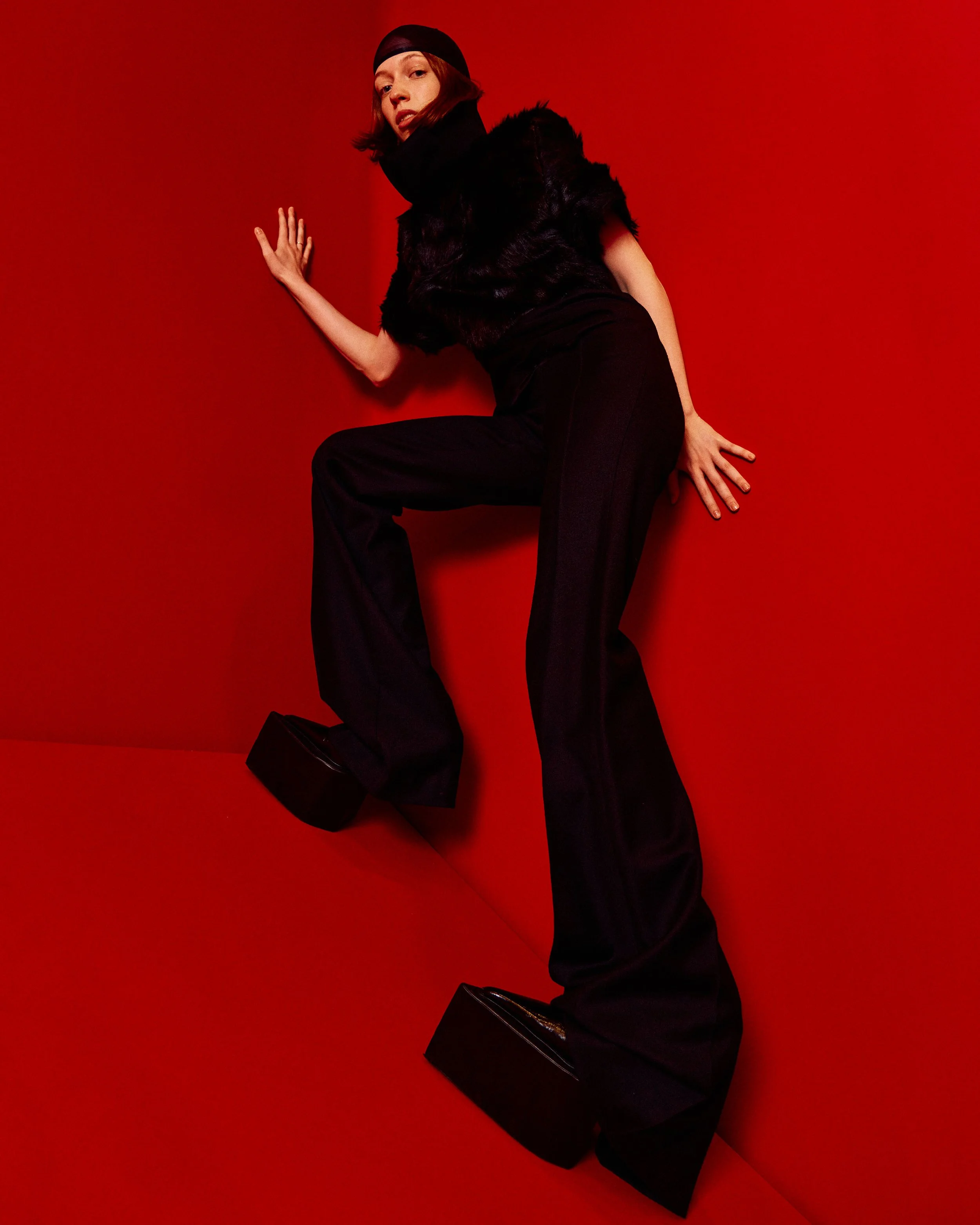 A woman with shoulder-length hair wearing a black headscarf, black furry vest, and loose black pants, is posing against a red background with her right hand on the wall and left hand on a black box.
