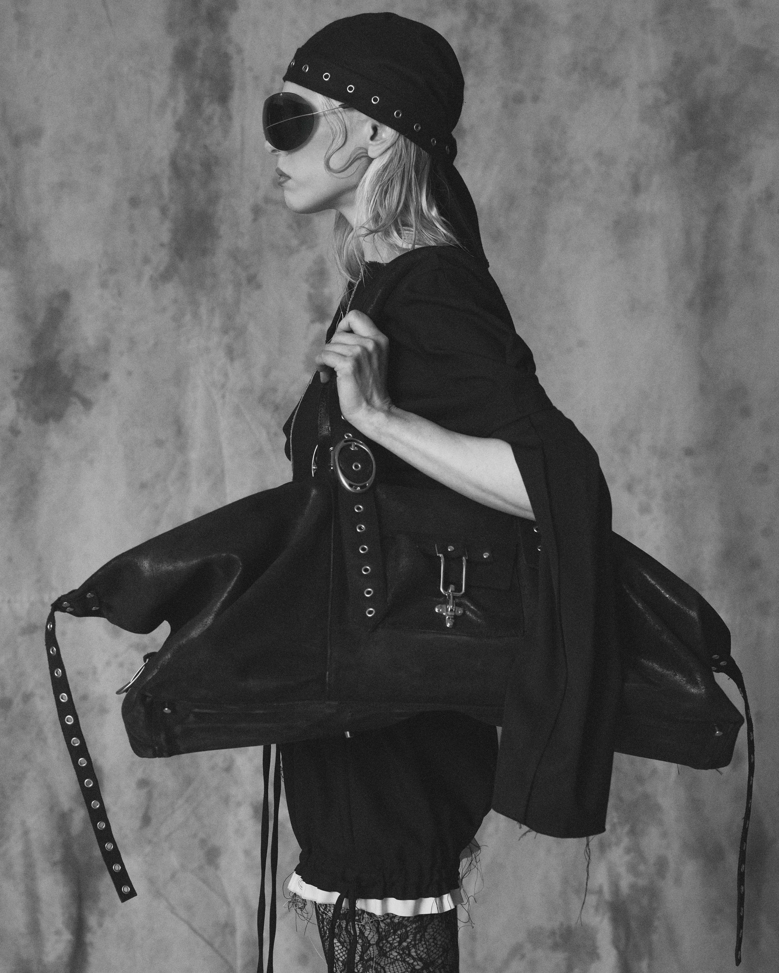 A black and white photograph of a woman in profile, wearing a black headscarf, large dark sunglasses, a black top, and fishnet stockings. She is holding a long, black, belt-like accessory with metal eyelets and rings over her shoulder, against a text
