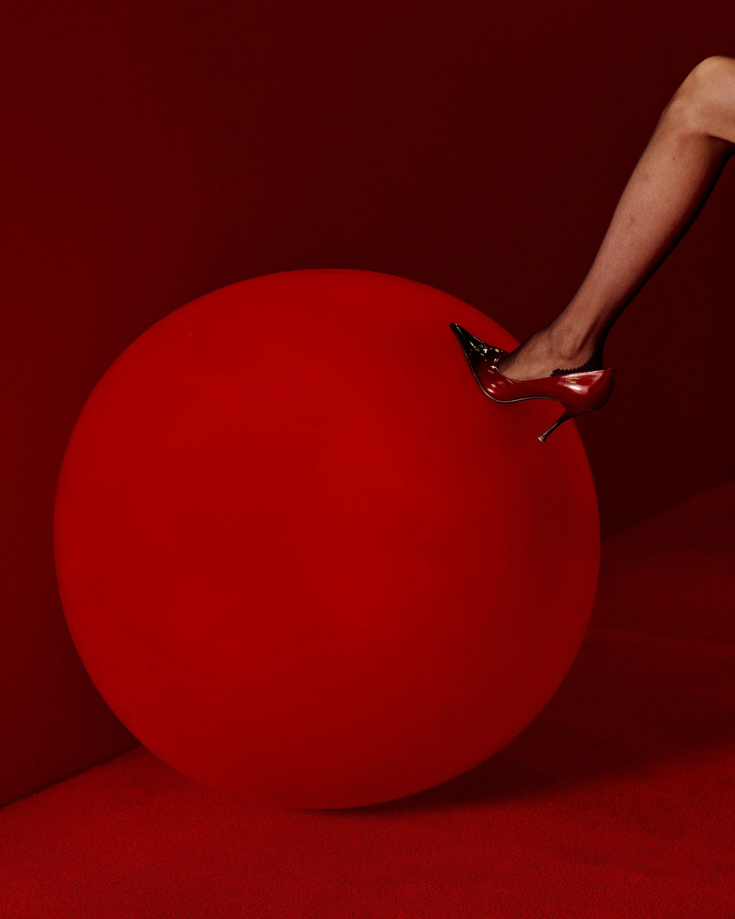 A person's leg wearing a shiny red high heel shoe, standing on a large red sphere with a red background.