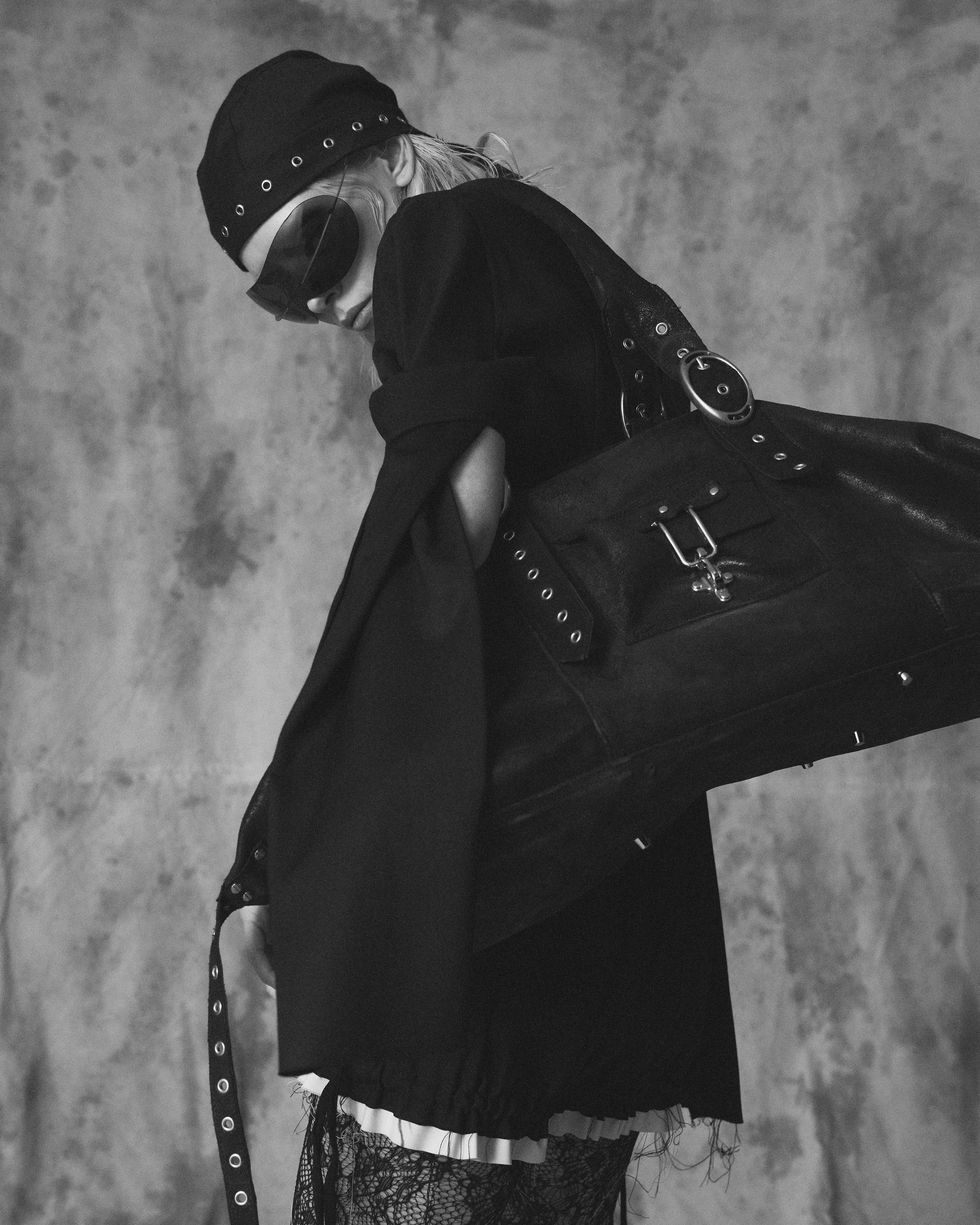 A person dressed in dark, layered clothing with a wide belt, wearing a cap and sunglasses, posing against a textured background.