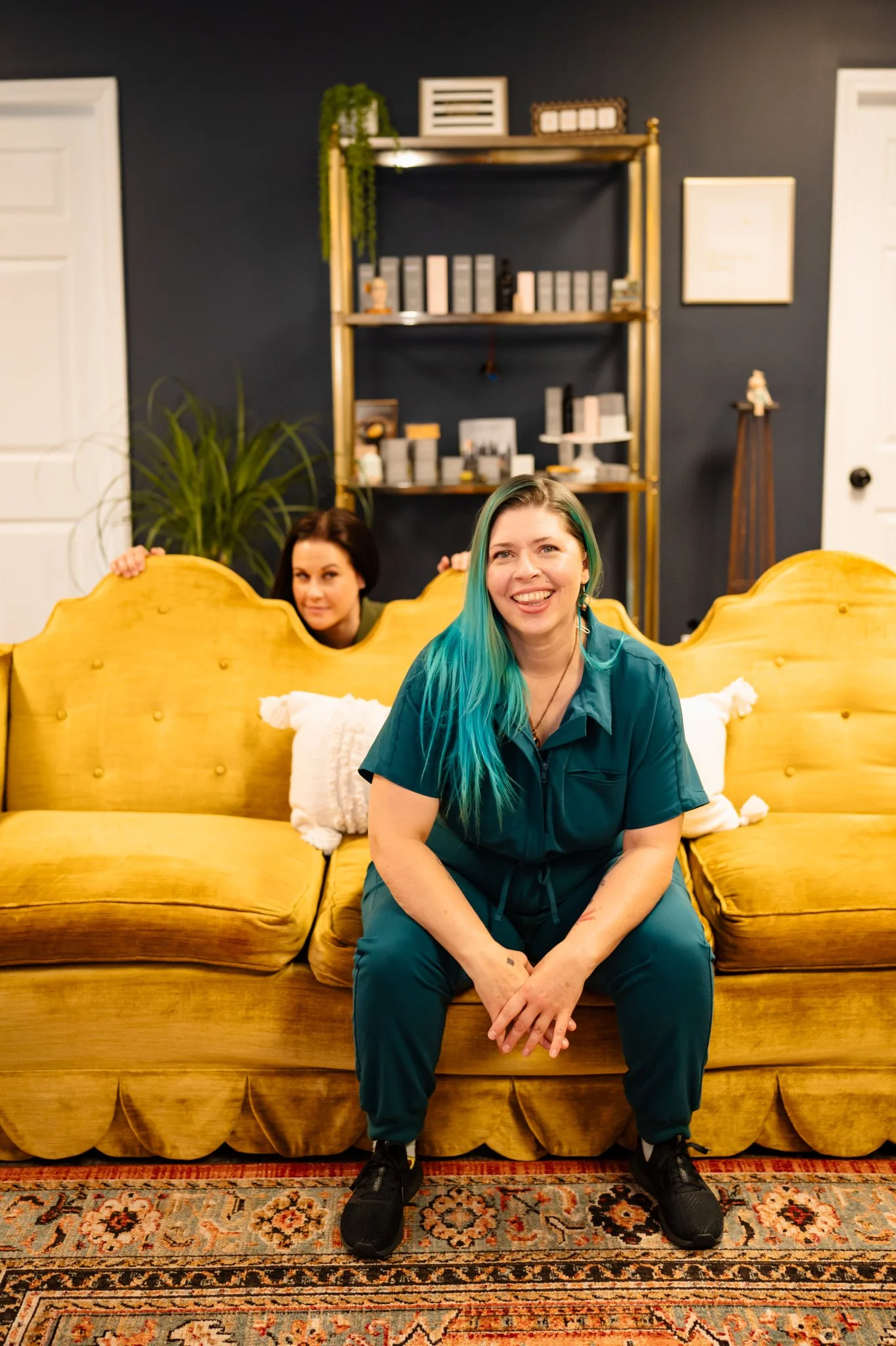 Two women, one with teal hair and the other with brown hair, are sitting on a yellow vintage-style couch in a room with dark blue walls. The woman with teal hair is in the foreground, smiling at the camera, while the woman with brown hair peeks from behind the couch. Behind them, there is a dark wall with a gold shelving unit holding decorative objects, and a large green plant on the left side.
