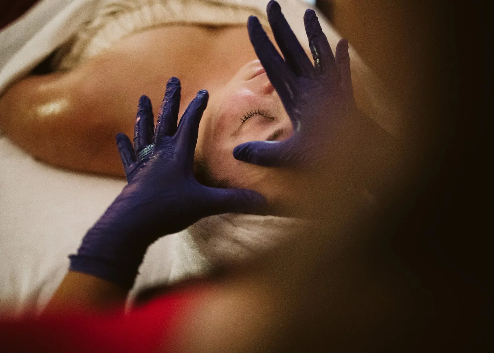 A person lying on a massage table with eyes closed, receiving a facial or skincare treatment from someone wearing blue gloves.