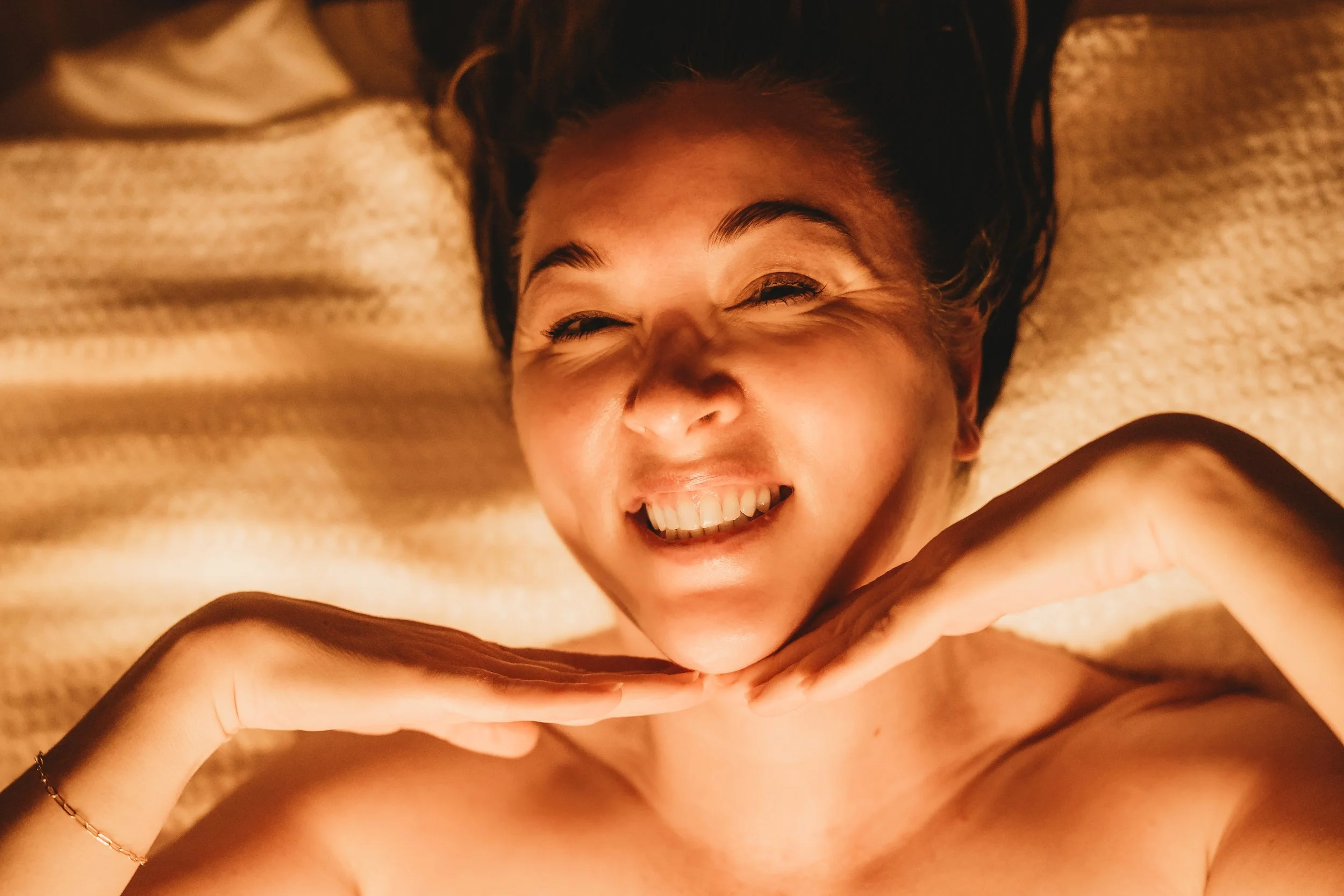Person lying on a blanket, smiling, with hand under chin, in warm lighting.