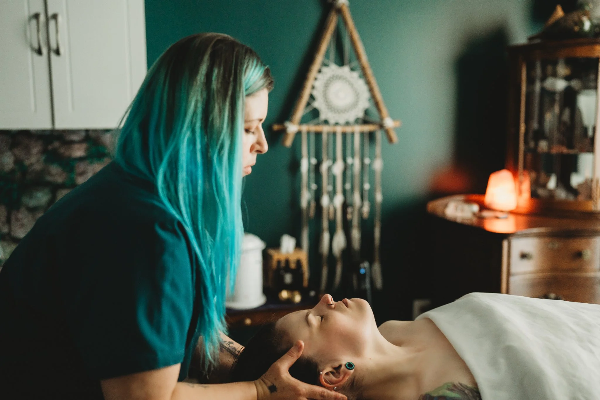 A woman with long, teal-colored hair is leaning over a person lying on a bed. The woman appears to be performing a massage or similar care, with her hands on the person's neck or shoulders. The room has a dark green wall, a dreamcatcher on the wall, and some furniture, including a lamp emitting an orange glow.