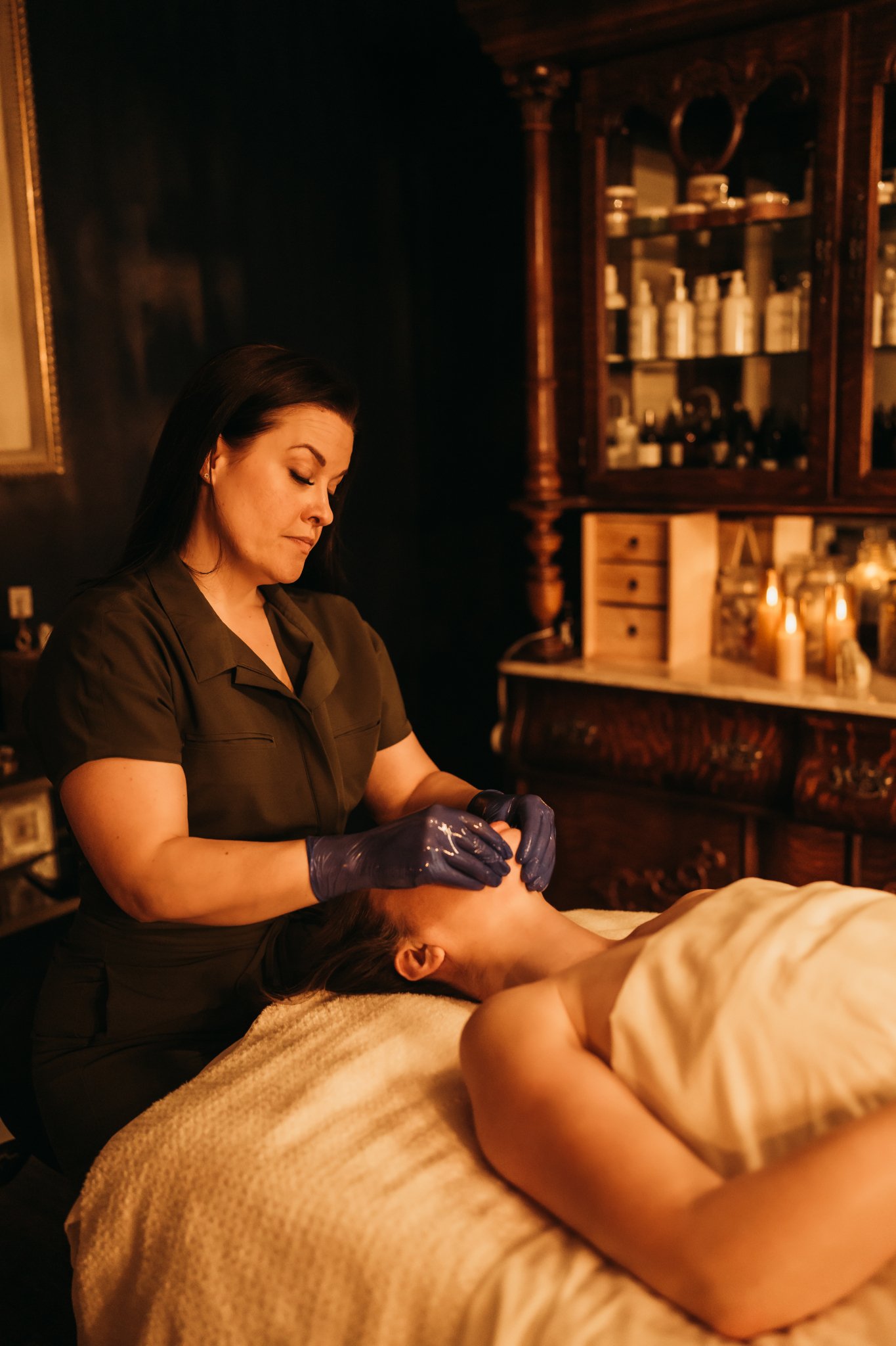 A woman receiving a facial massage with a therapist wearing black gloves in a cozy, dimly lit room.