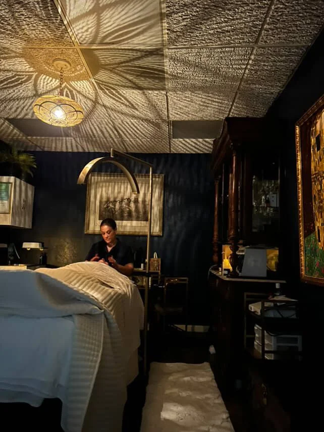A person standing near a bed in a dimly lit room, illuminated by a decorative ceiling light casting shadows on the ceiling. The room has dark walls and ornate furniture, including a wooden cabinet and framed artwork.