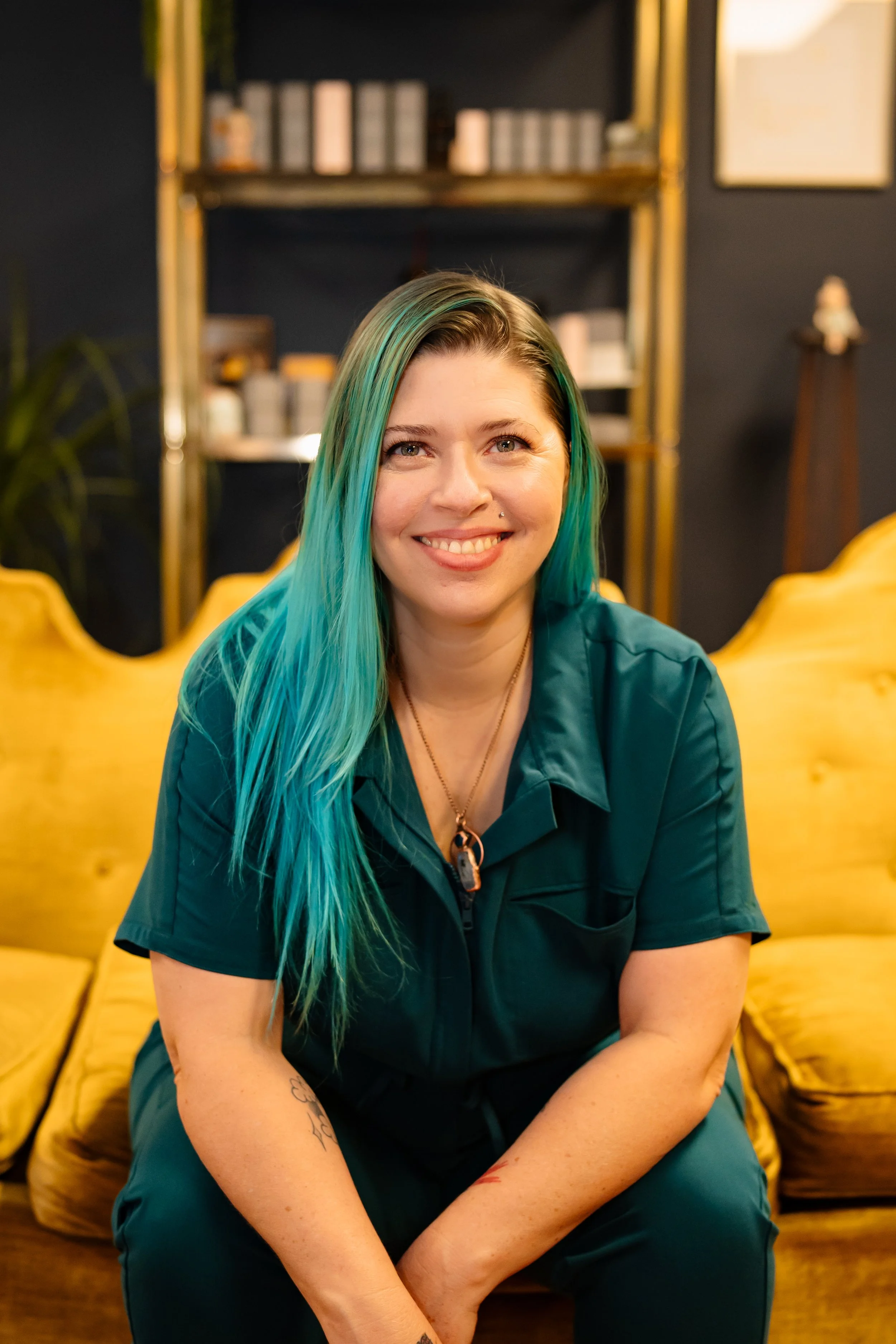 A woman with long turquoise hair smiling, sitting on a yellow sofa in a room with dark walls and shelves in the background.