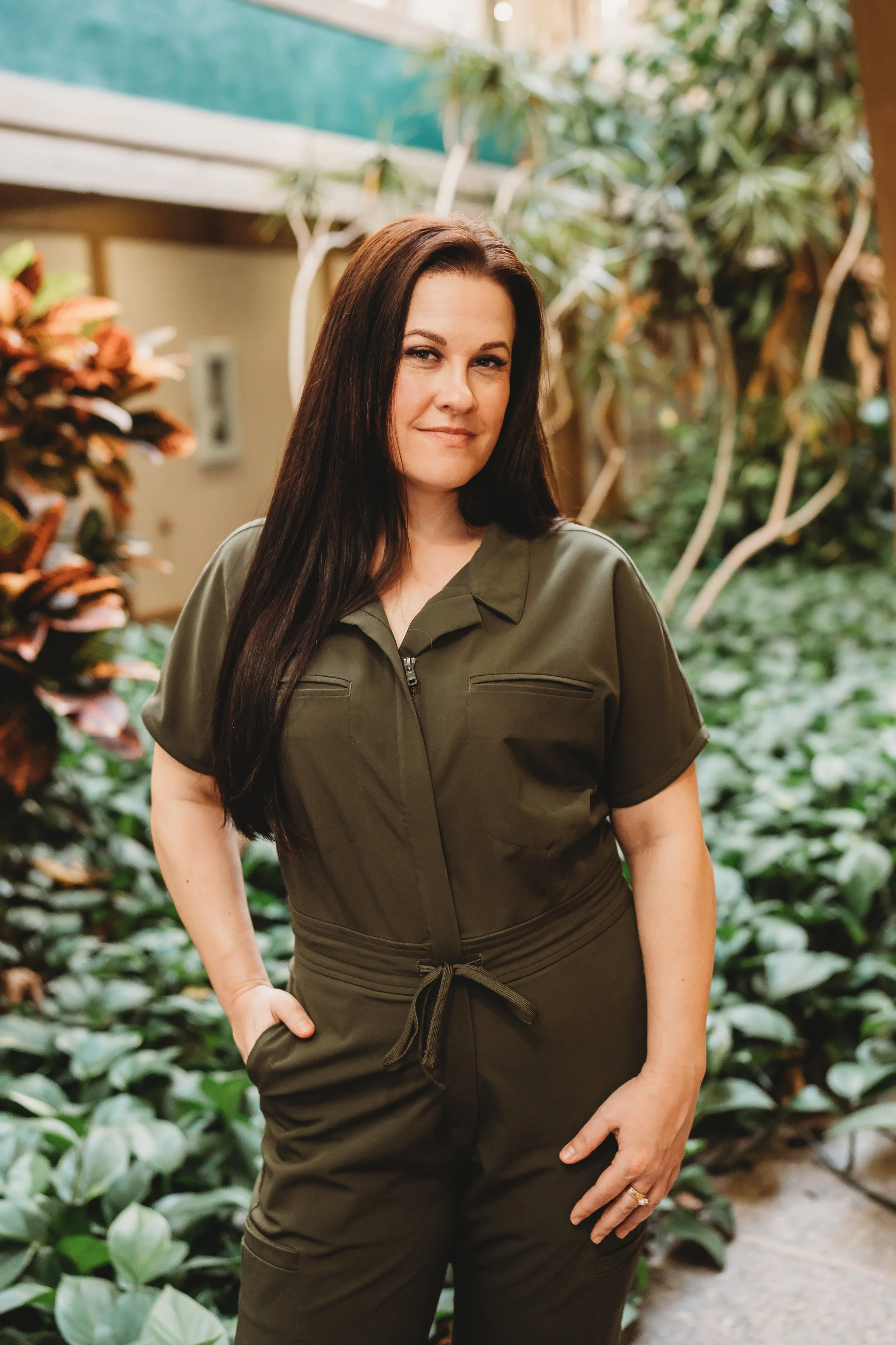 A woman with long brown hair standing in a lush indoor garden, wearing a green zip-up jumpsuit with her left hand on her hip, smiling subtly at the camera.