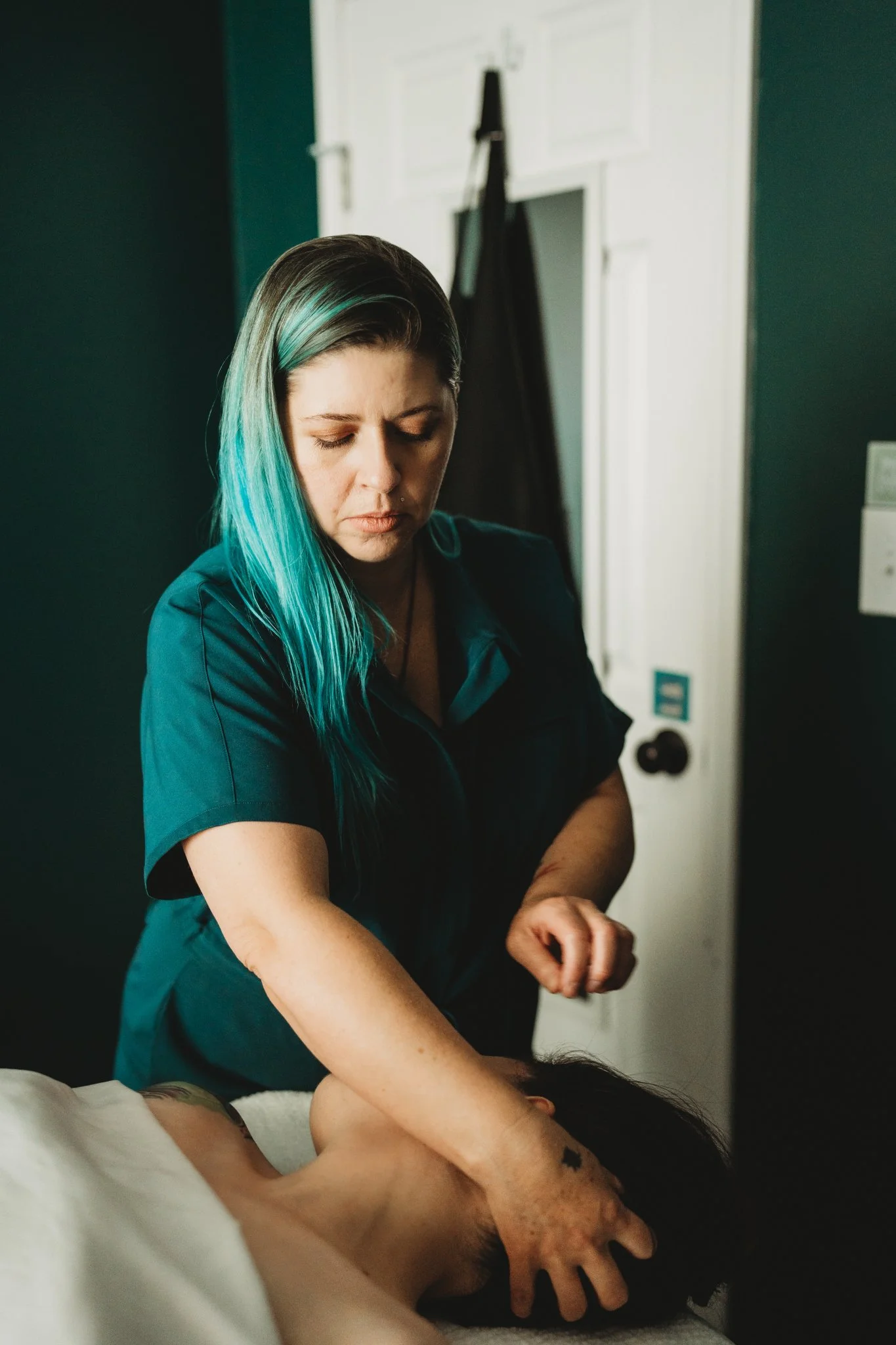 A woman with teal and black hair giving a massage to a person lying face down on a bed, with a black wall and white door in the background.