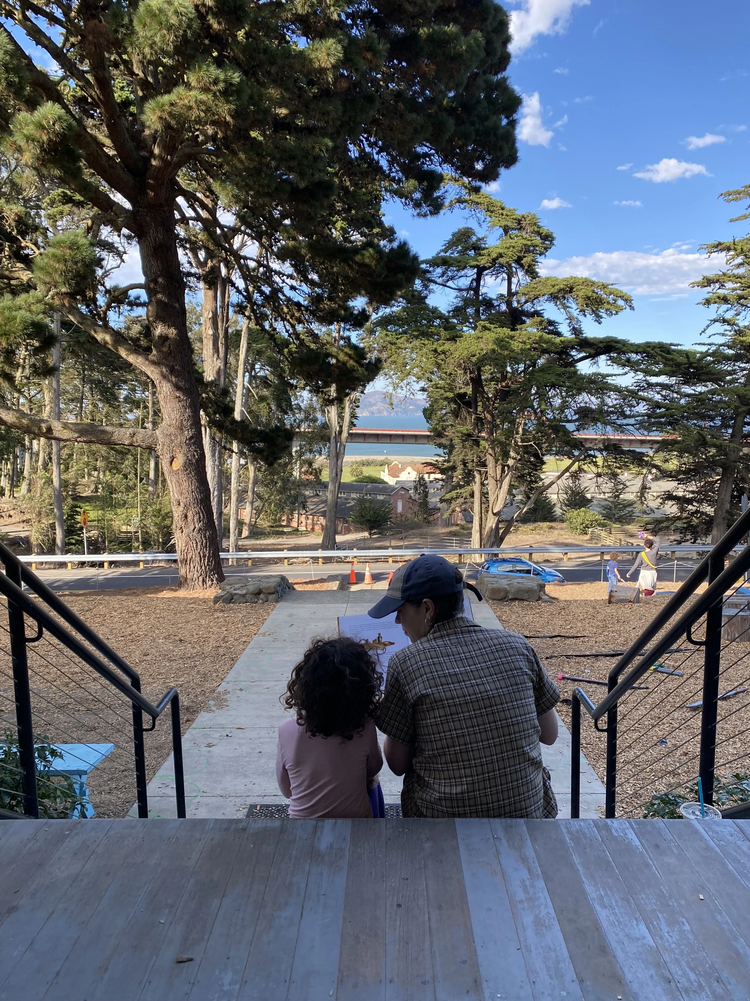 Two people sitting on stairs, a man and a young girl, outdoors on a sunny day with tall trees and a road in the background.
