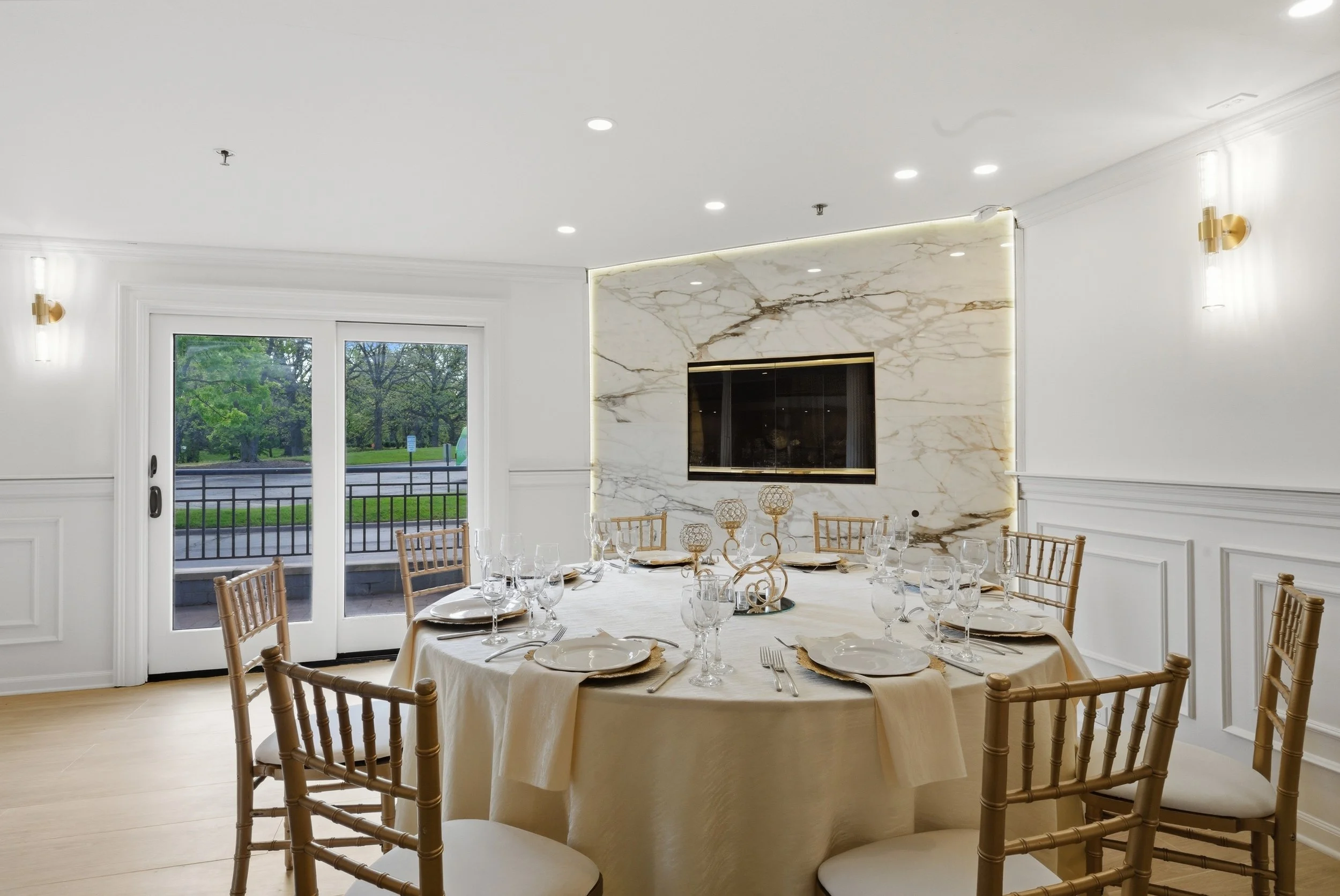 A dining room with a round table set with white tablecloth, glassware, and gold-colored chairs, with a marble fireplace, large windows, and lush green trees visible outside.