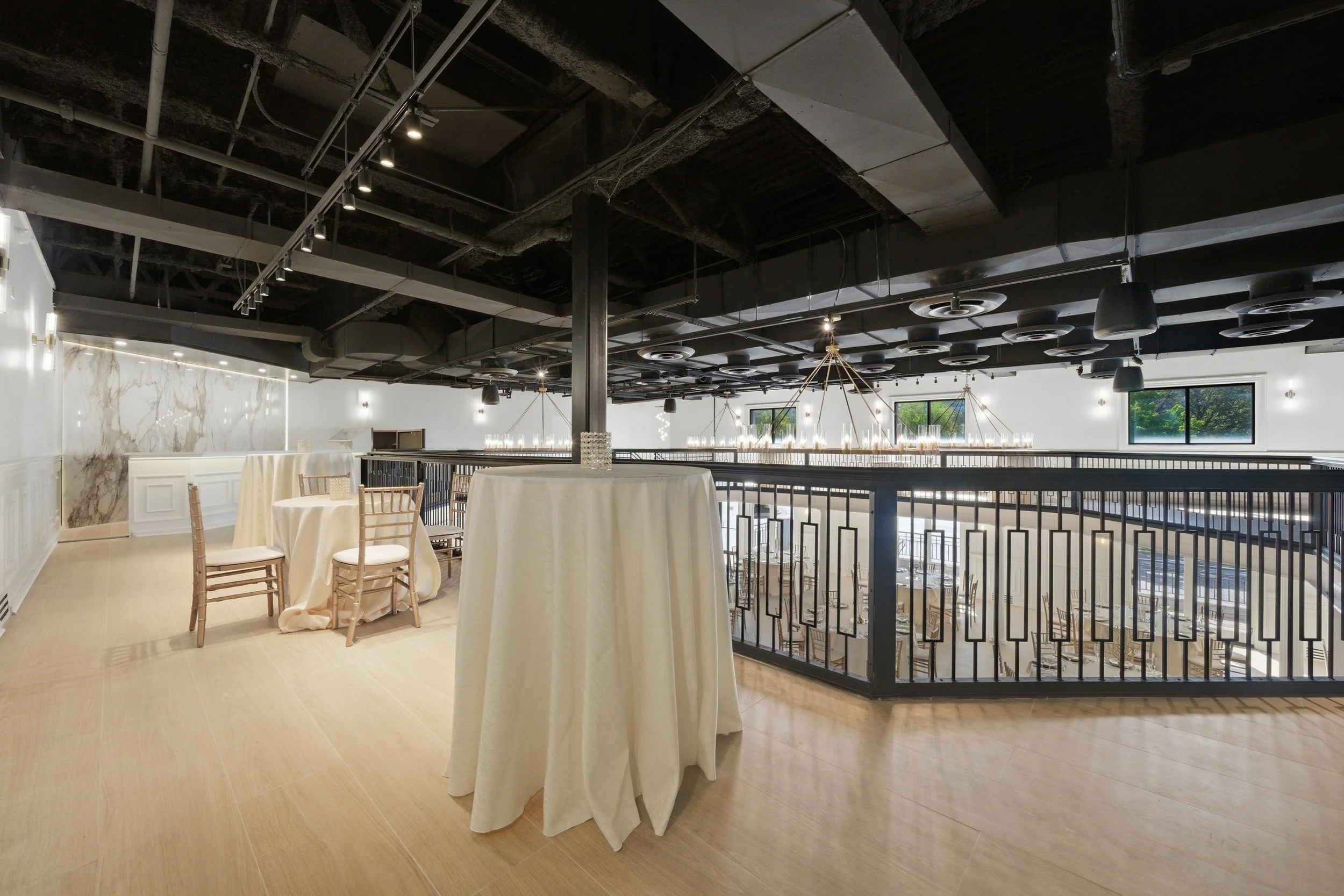 Elegant event space with round tables covered in white tablecloths, set for a formal gathering. The room features a modern black ceiling with circular air vents and hanging lights, large windows with natural light, and a decorative metal railing on t
