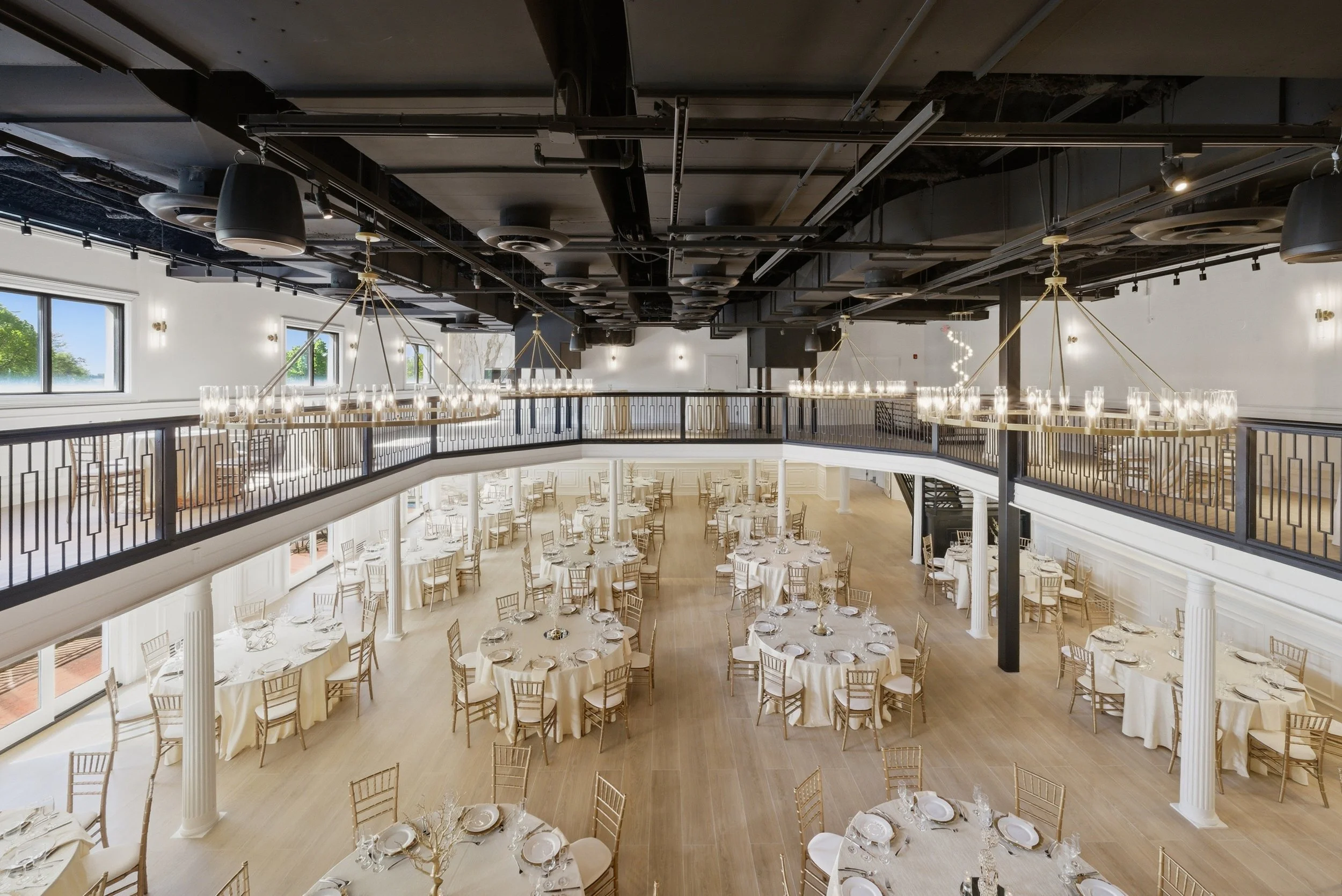 Elegant event space with round tables covered in white tablecloths and silverware, set up for a formal dinner. A mezzanine level with a black railing overlooks the area. Large chandeliers with candles hang from a high ceiling with black exposed ductw