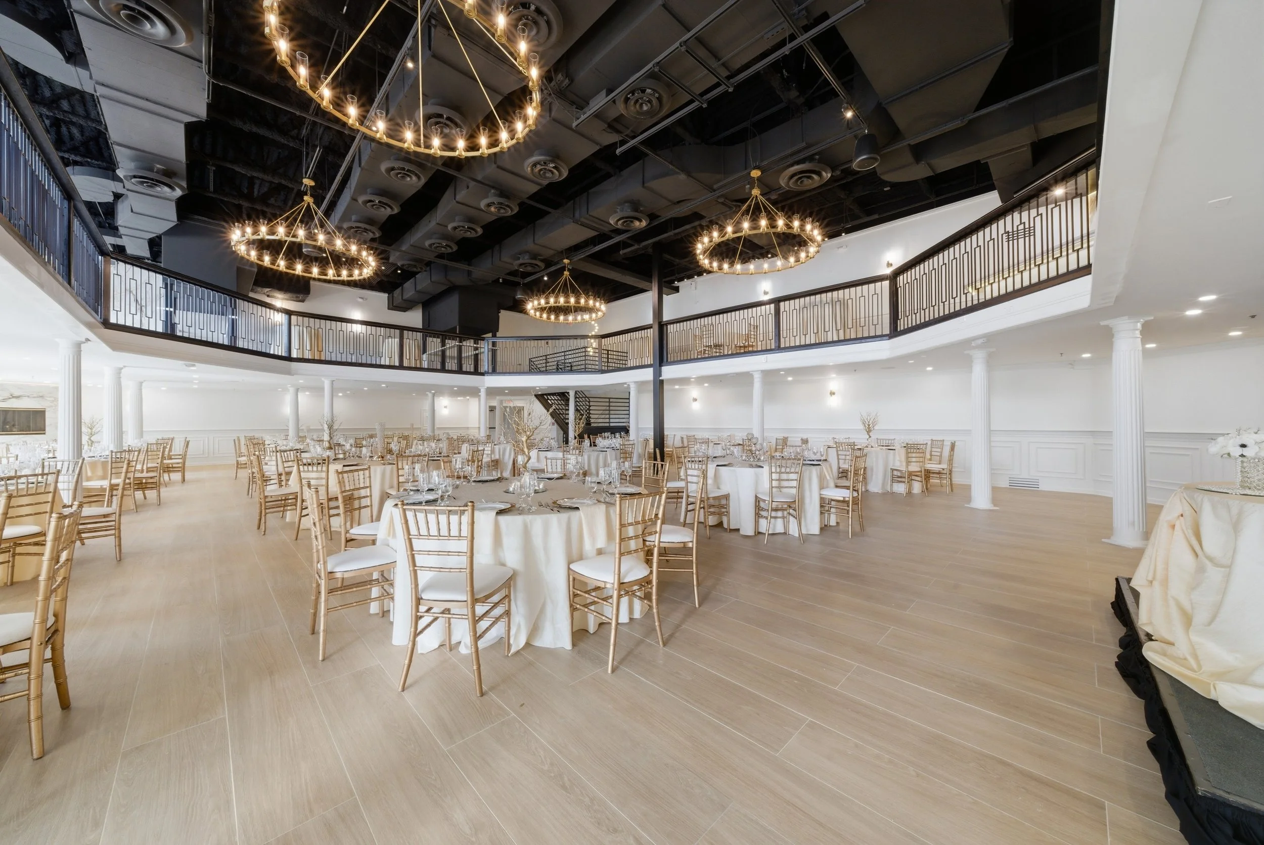 Empty banquet hall with round tables covered in beige tablecloths, set with glassware and white chairs, white walls with decorative moldings, and a second-floor balcony with black railing. Three large, circular chandeliers with lights hang from the d