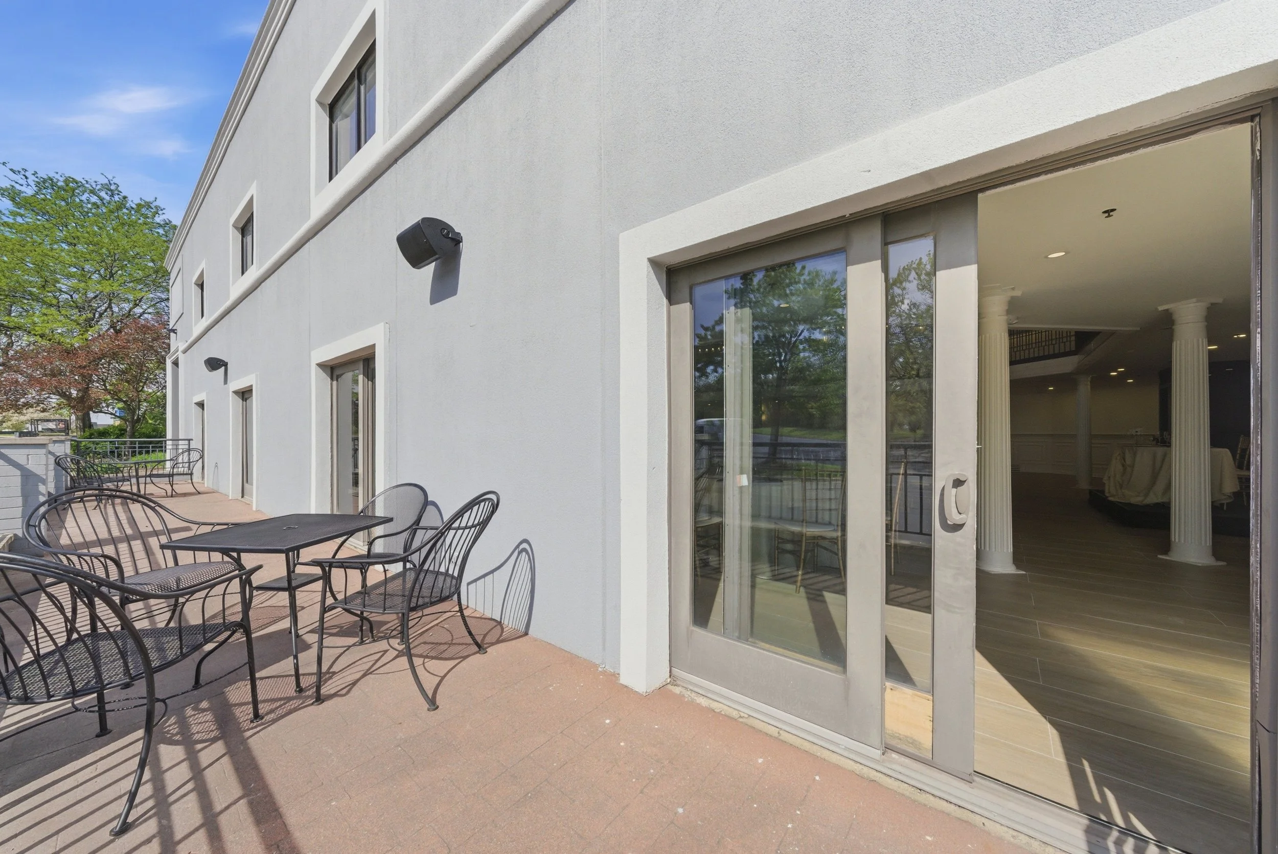 Outdoor patio area with black metal chairs and tables, adjacent to a white building with large sliding glass doors and windows, and a view of trees and a clear blue sky.