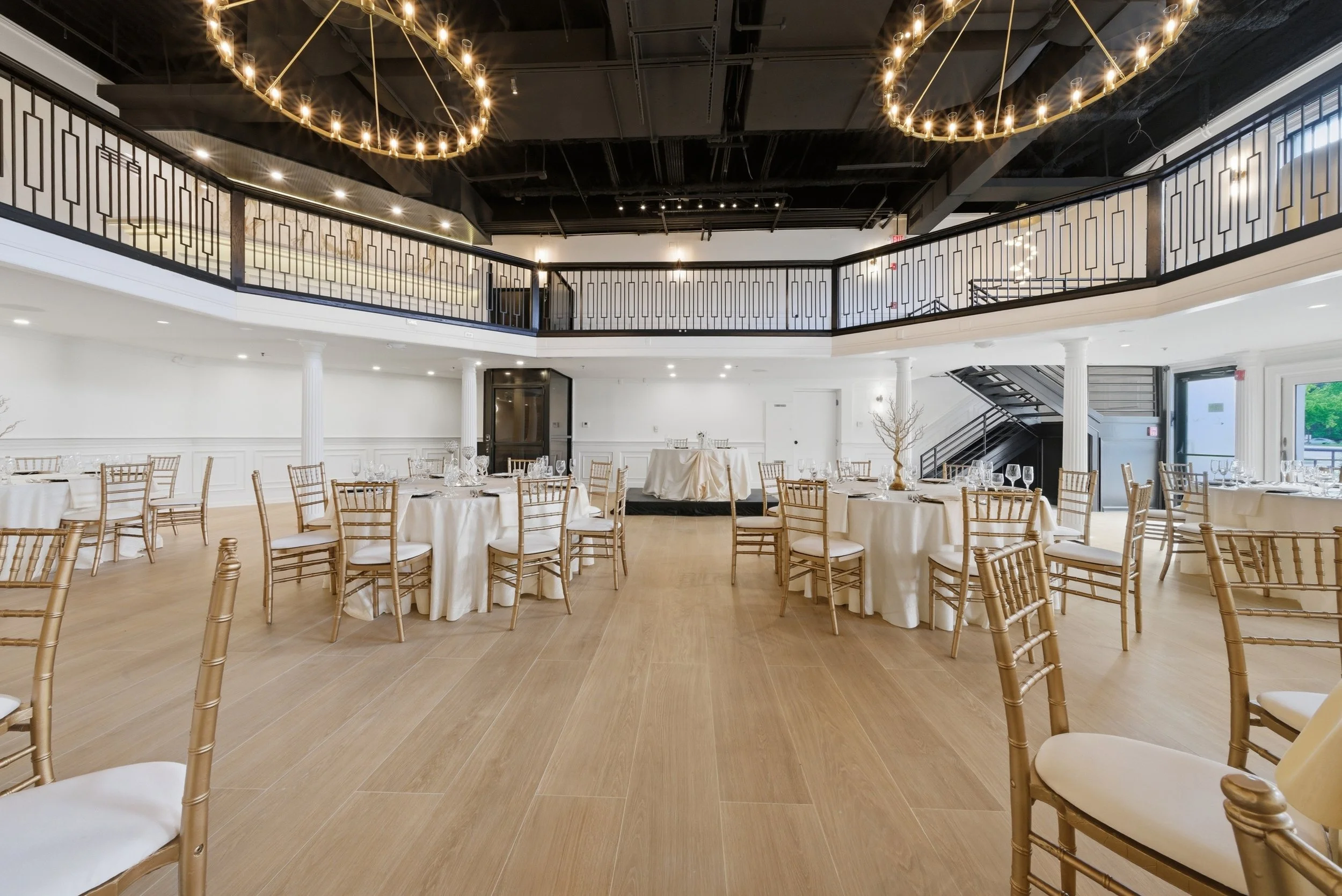 Elegant banquet hall decorated with round tables covered in white tablecloths, gold chairs, and glassware. The ceiling features black beams and two large gold chandeliers with multiple light bulbs. White columns support the upper balcony, which has a