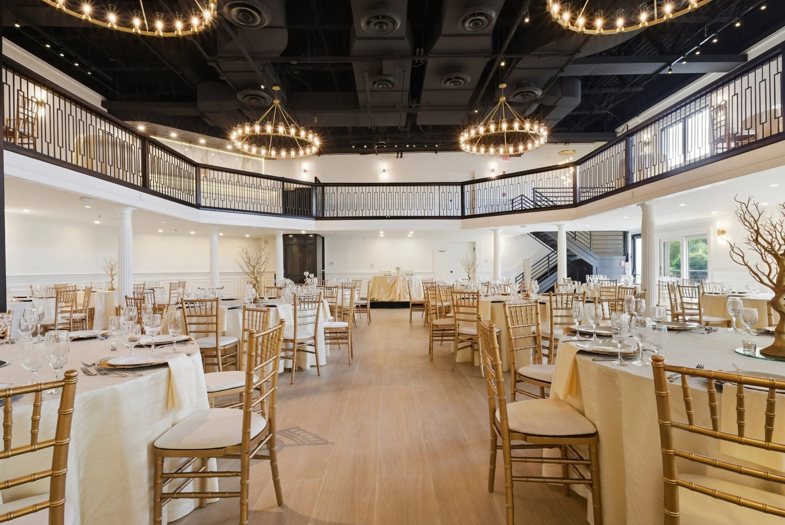 Elegant banquet hall with round tables covered in white tablecloths, gold Chiavari chairs, and chandeliers hanging from the black ceiling. The room features a second-floor balcony with black railings, white columns, and a staircase, with large window