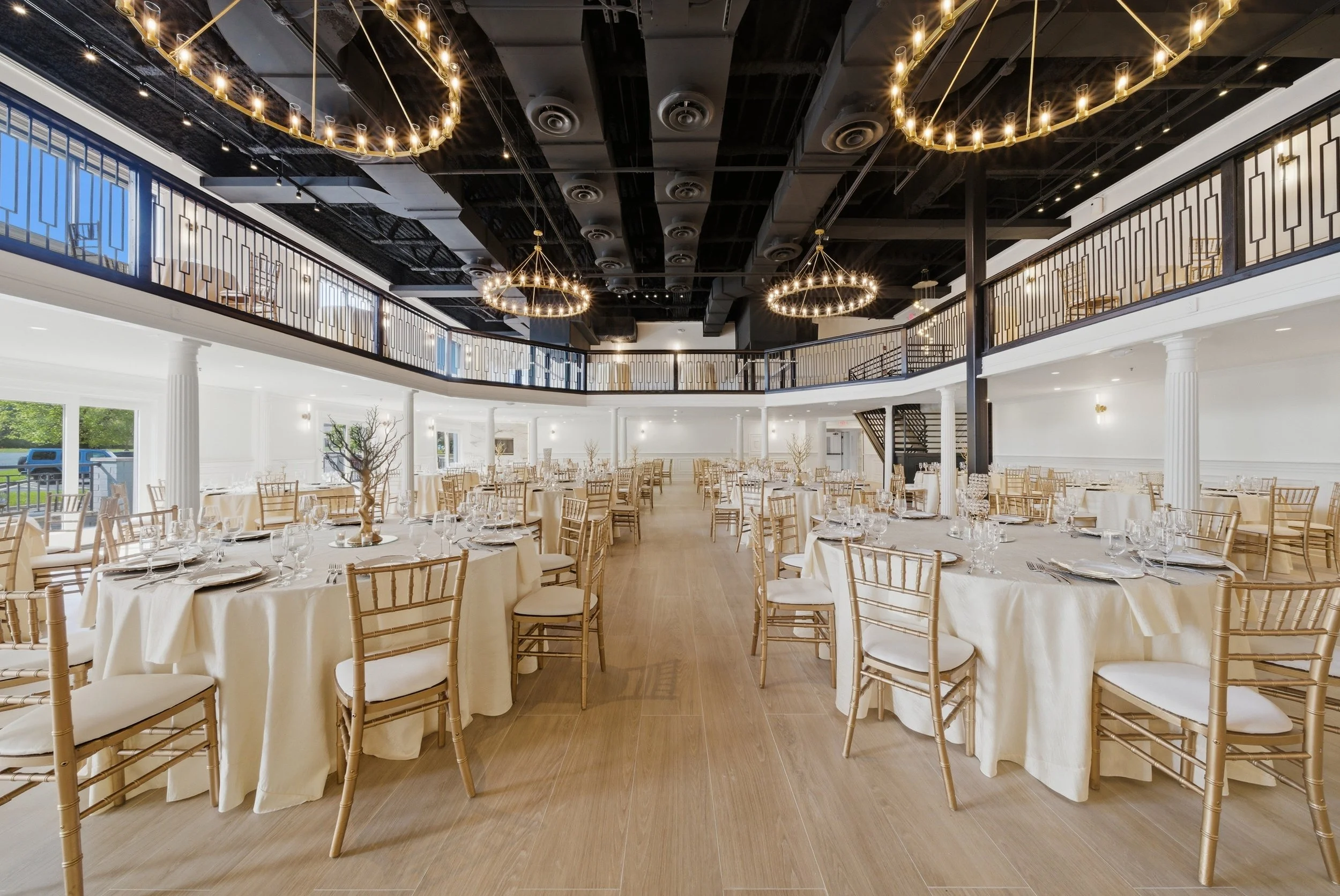 Elegant banquet hall decorated for an event with round tables covered in white tablecloths and surrounded by gold chairs. The hall features an open second-floor balcony with black railing, large windows, and modern chandelier lighting fixtures hangin