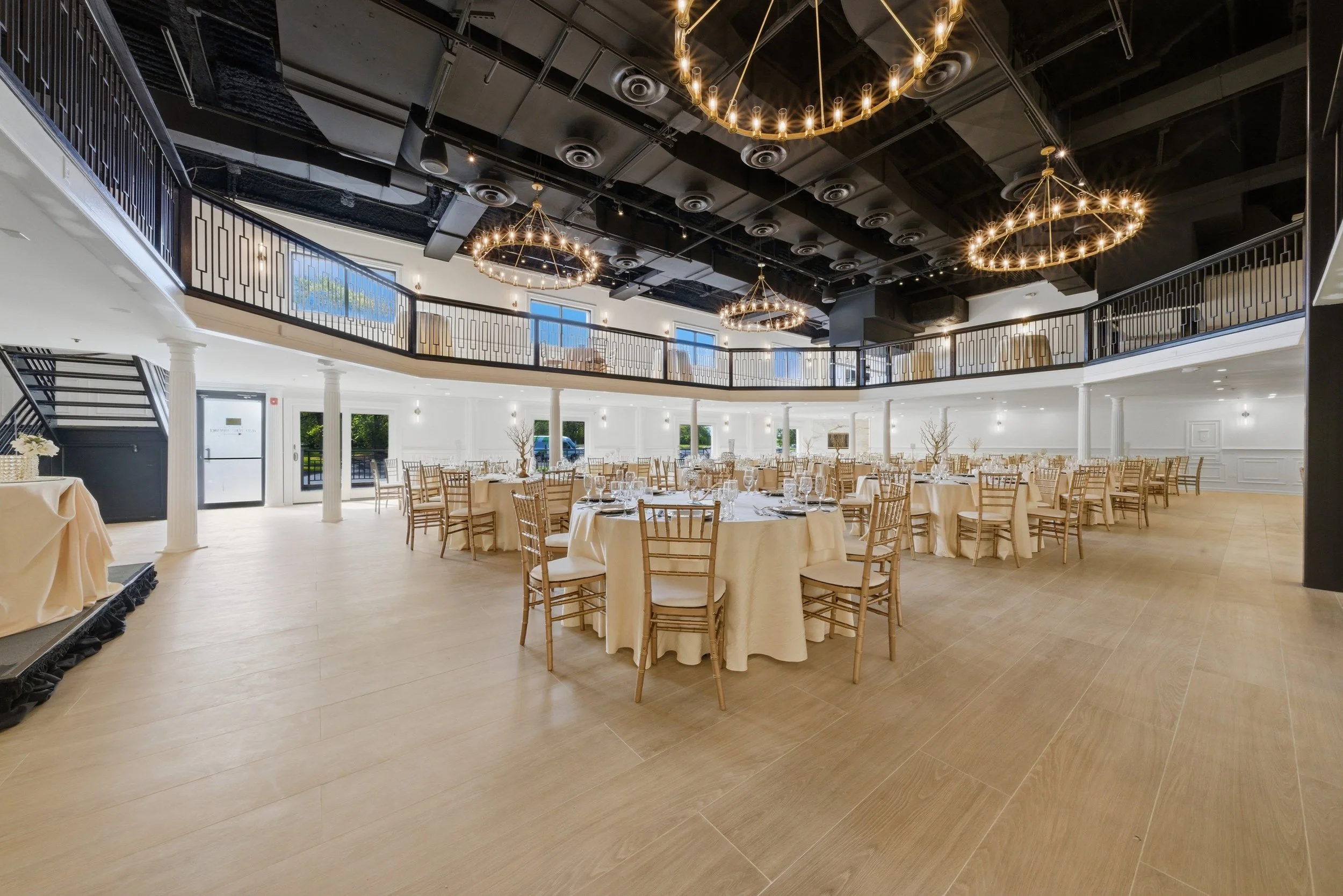 Elegant banquet hall with round tables draped in white tablecloths, set with glassware and silverware, beige chairs, and tall windows with natural light, featuring black chandeliers hanging from a black ceiling.