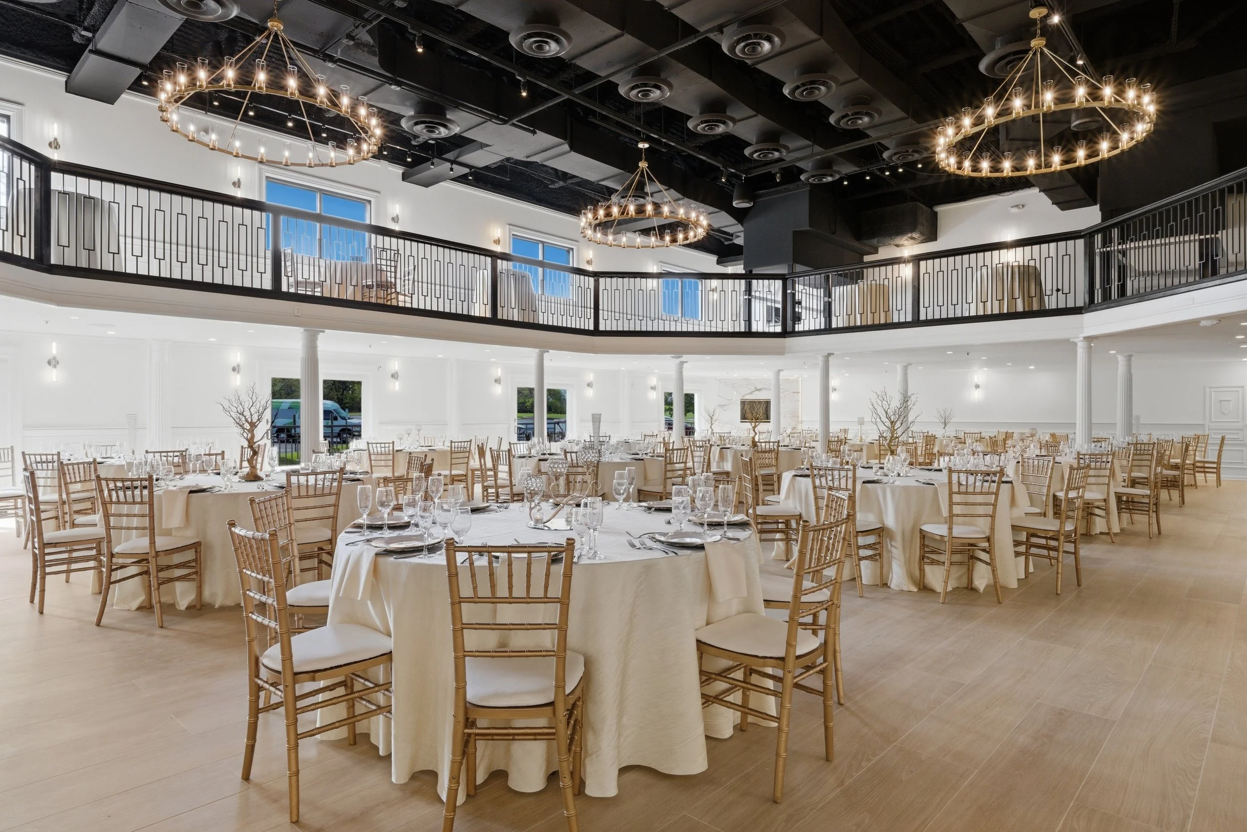 Elegant event hall with round tables covered in white tablecloths, gold Chiavari chairs, and glassware set for a formal gathering. The space features high ceilings, large windows, and a mezzanine with black railings. Suspended circular chandeliers wi